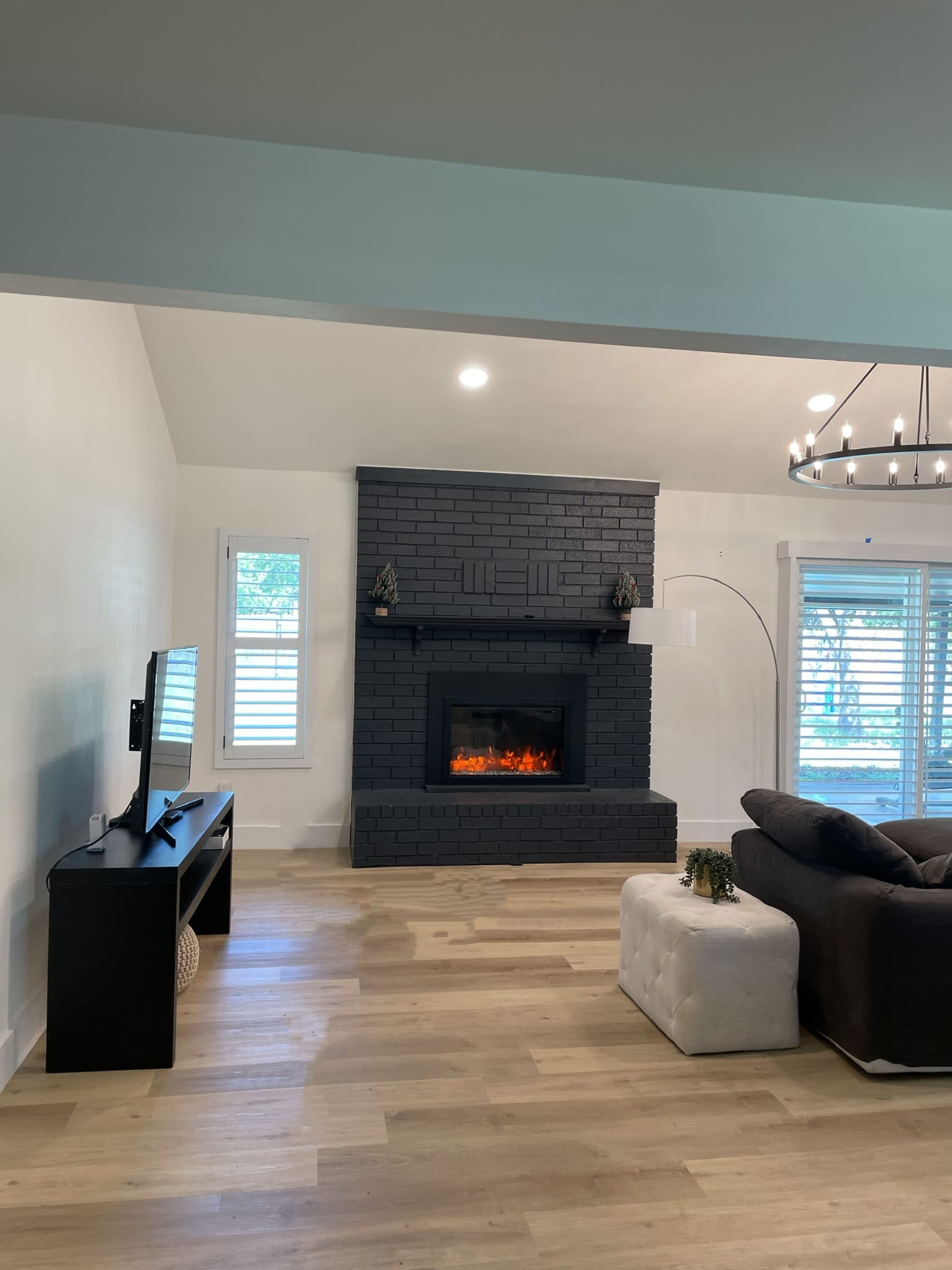 The living room features a black-brick fireplace, a television stand, a sectional sofa, and a white ottoman on a wooden floor.