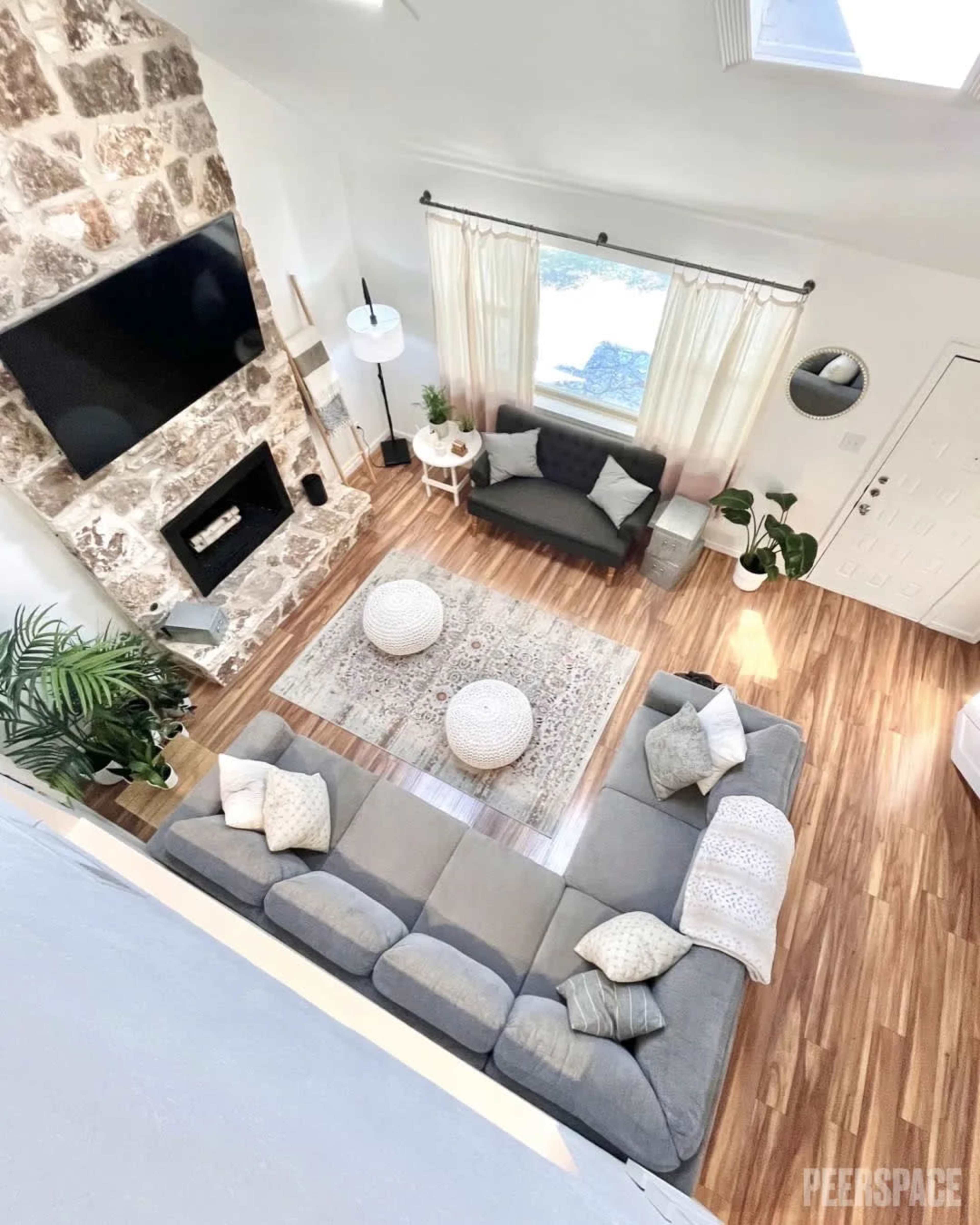 A spacious living room features a large gray sectional sofa, a stone fireplace, and a patterned area rug, with large windows allowing natural light to fill the space.