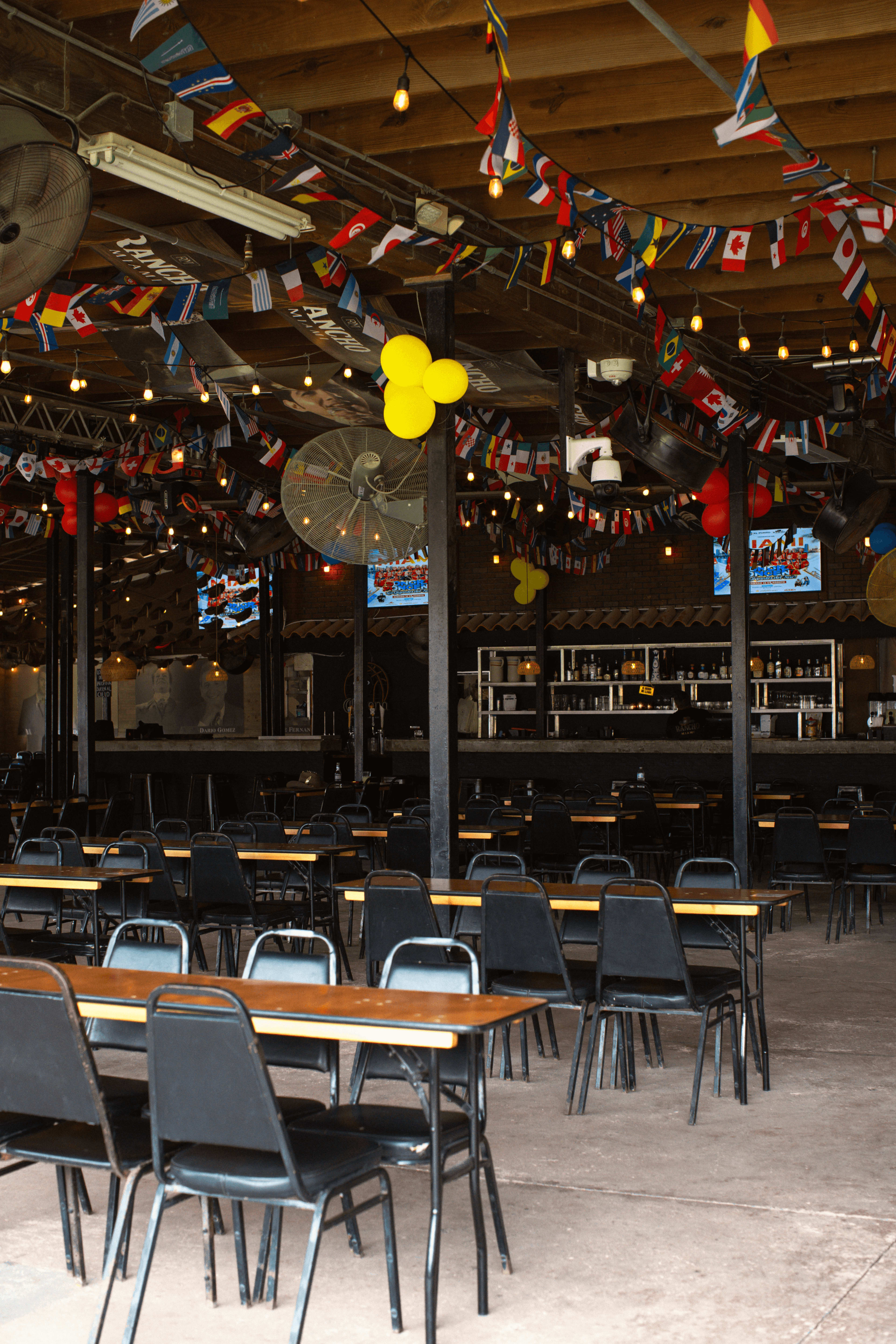 An indoor dining area showcases rows of tables and chairs beneath colorful international flags and string lights, with a bar visible in the background.