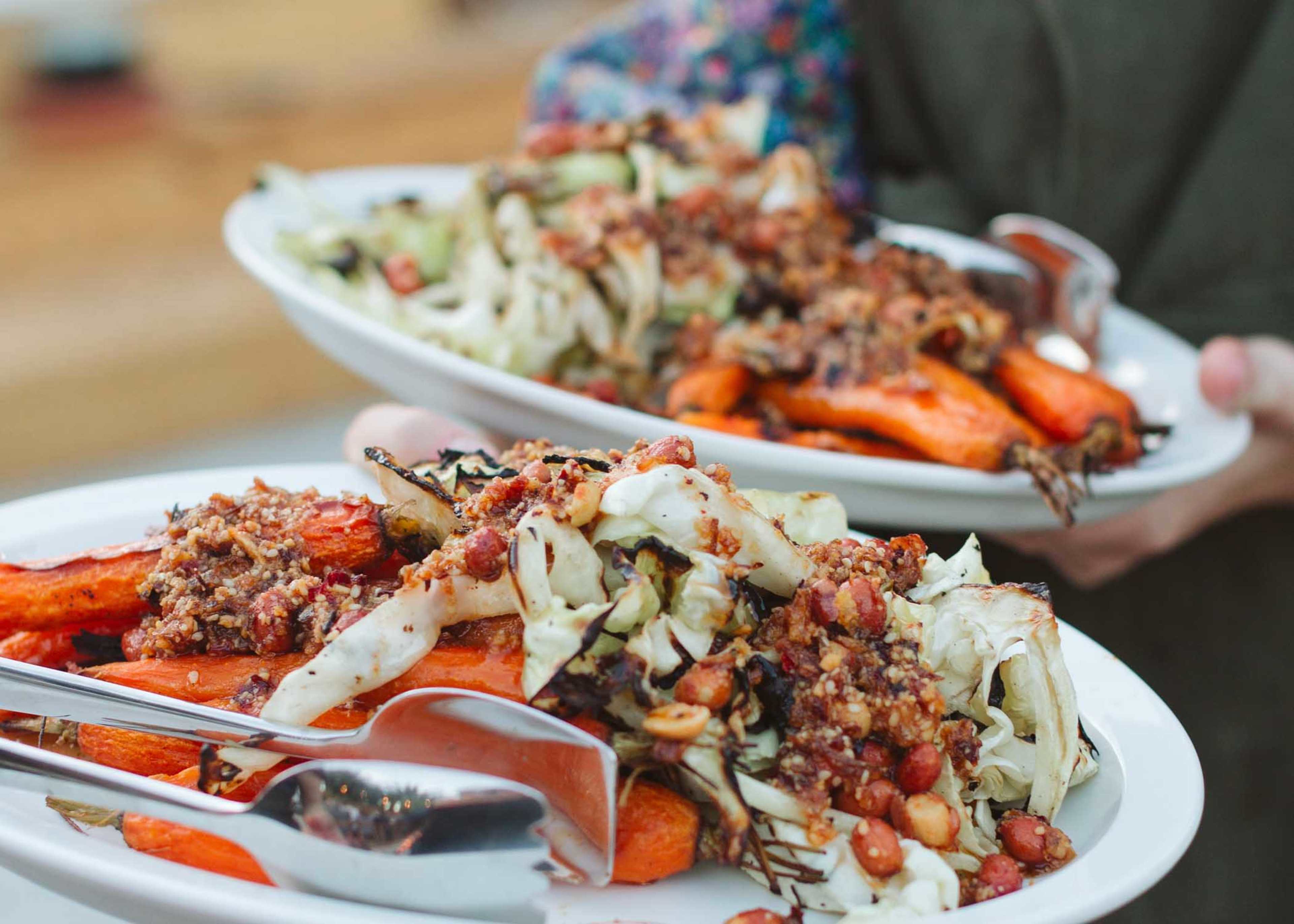 Two platters of roasted carrots and cabbage topped with a mixture of nuts and spices are being held by a person.