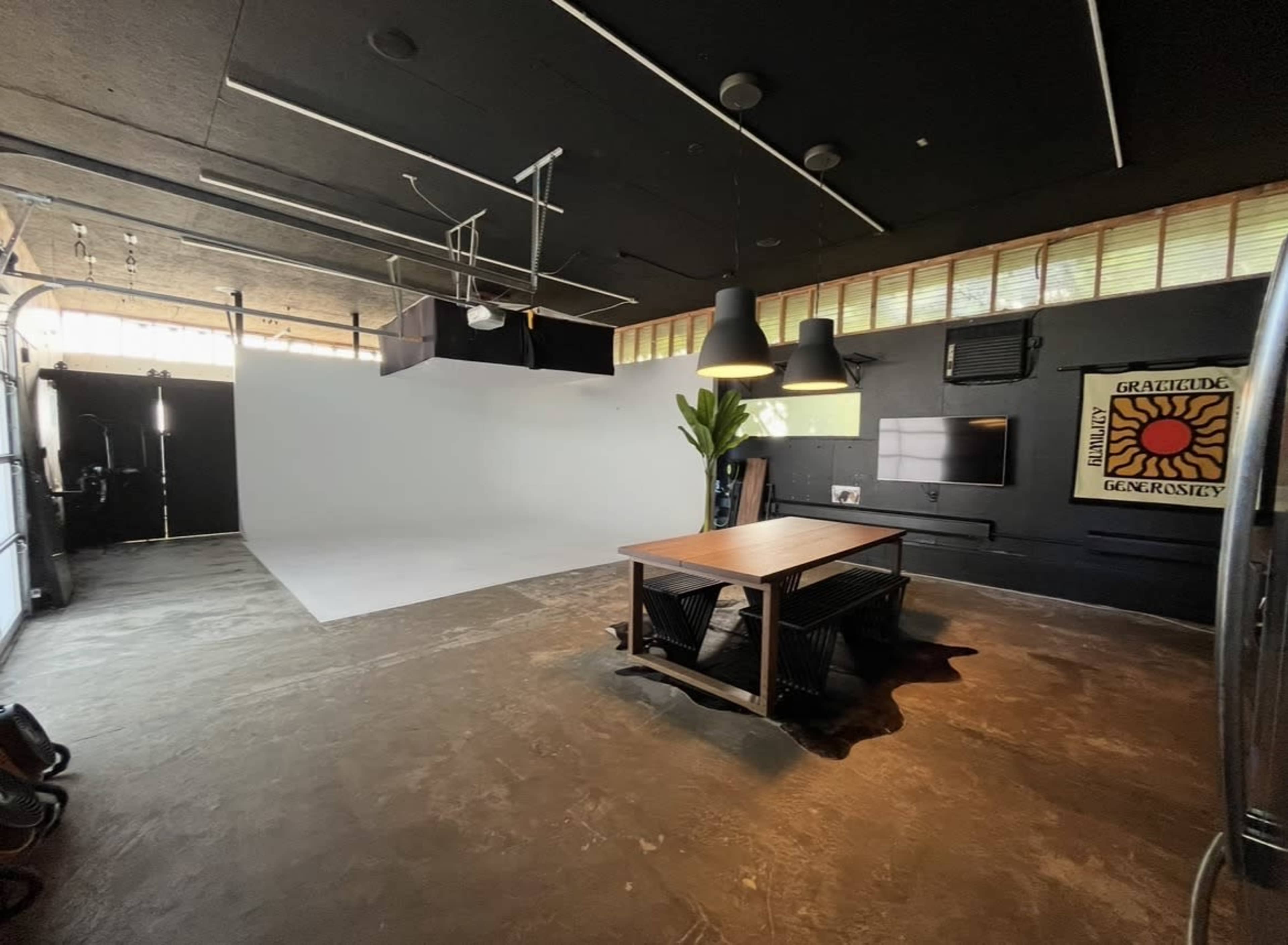 The image depicts a photography studio featuring a large white backdrop, a wooden table at the center, and various lighting fixtures hanging overhead.
