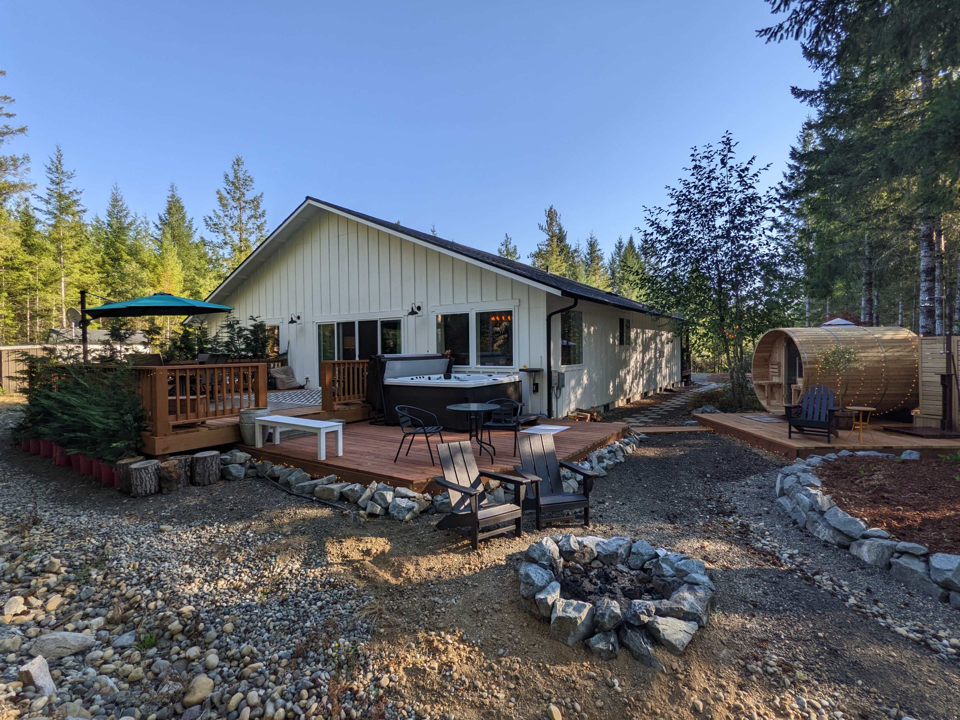 A modern cabin with a wooden deck, outdoor seating, and a hot tub surrounded by trees and a gravel pathway.