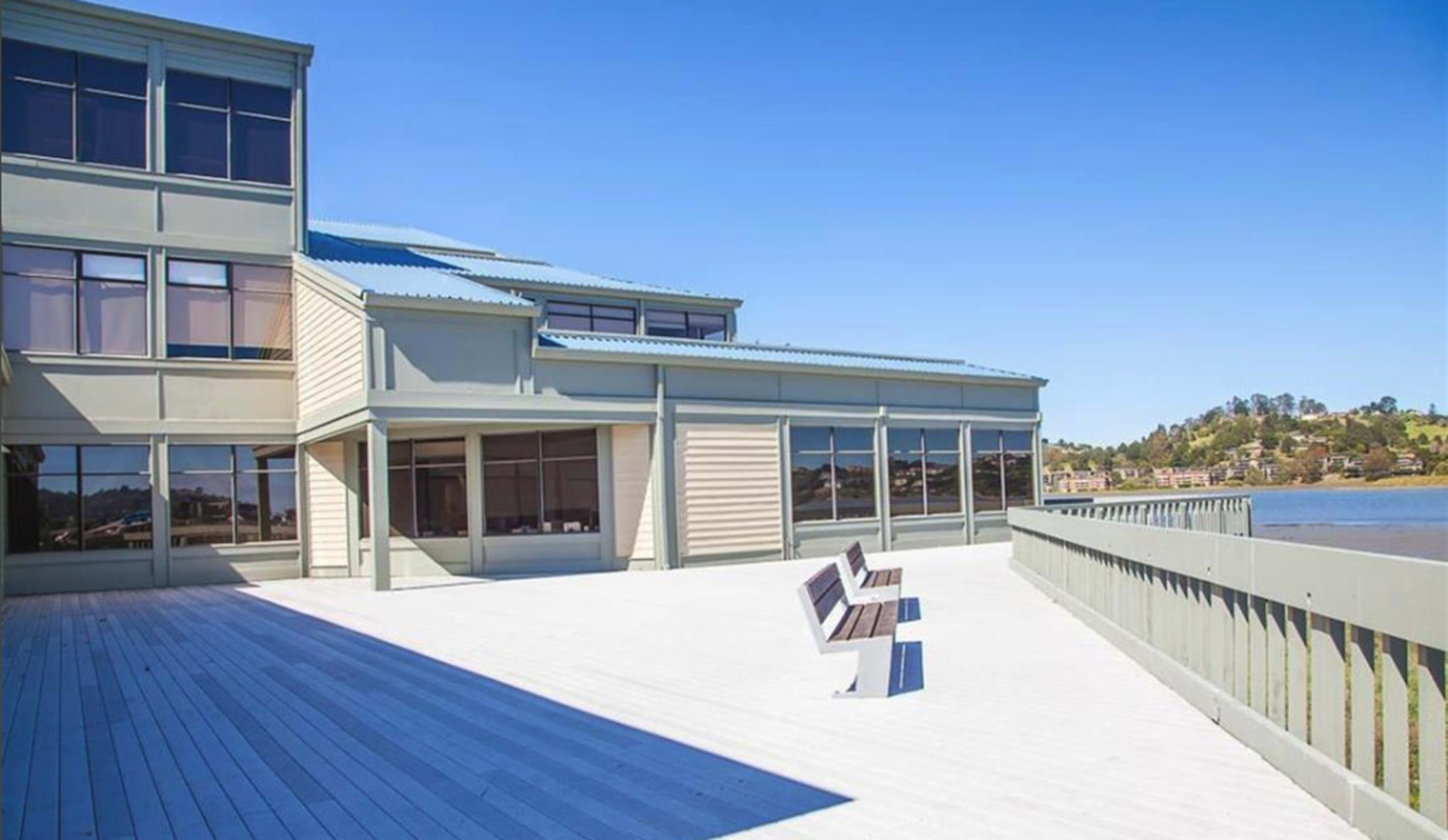 A deck with two benches overlooks a body of water, adjacent to a multi-story building with large windows.