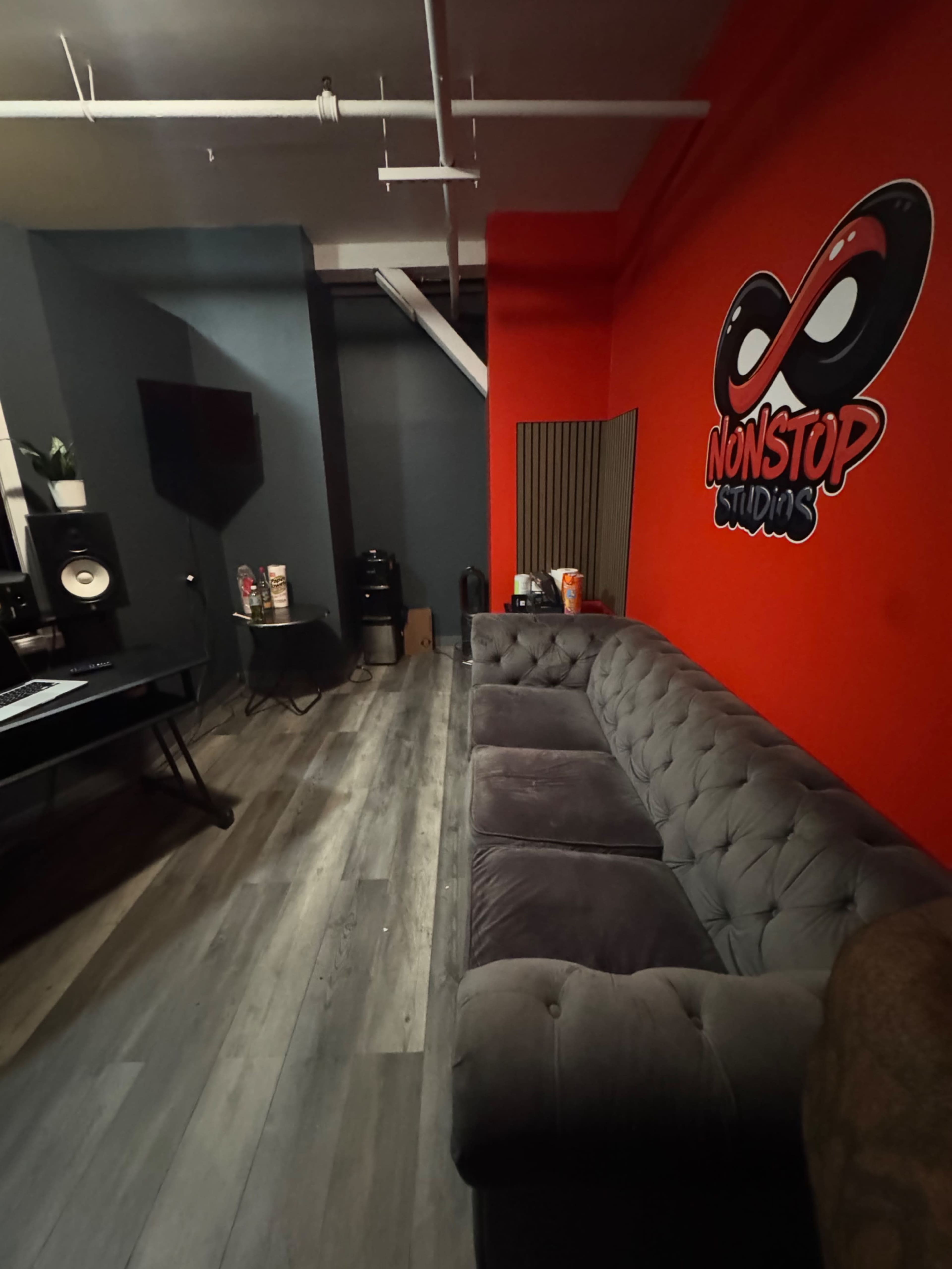 The image shows a cozy music studio with a tufted gray sofa, a red accent wall featuring the "NONSTOP STUDIOS" logo, and various music equipment arranged in the space.
