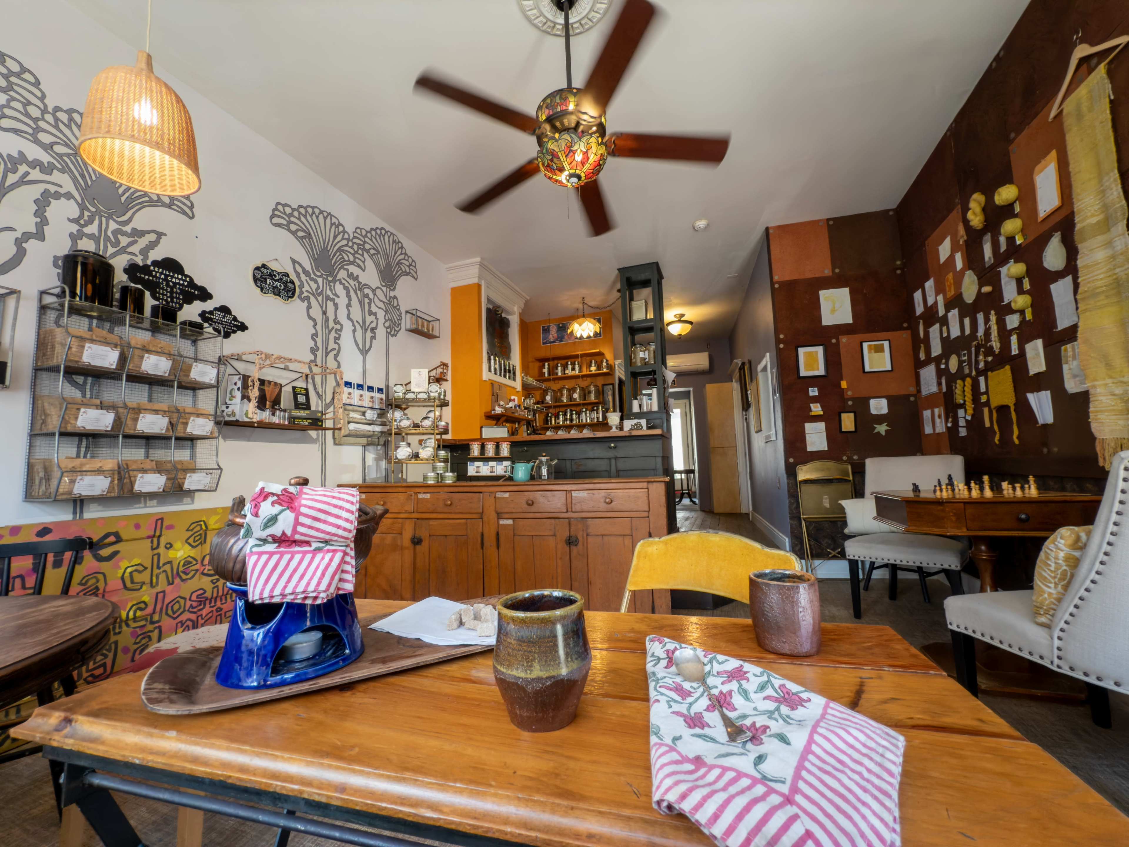 The image shows a cozy café with wooden furniture, a colorful wall mural, shelves displaying various items, and a bar area in the background.