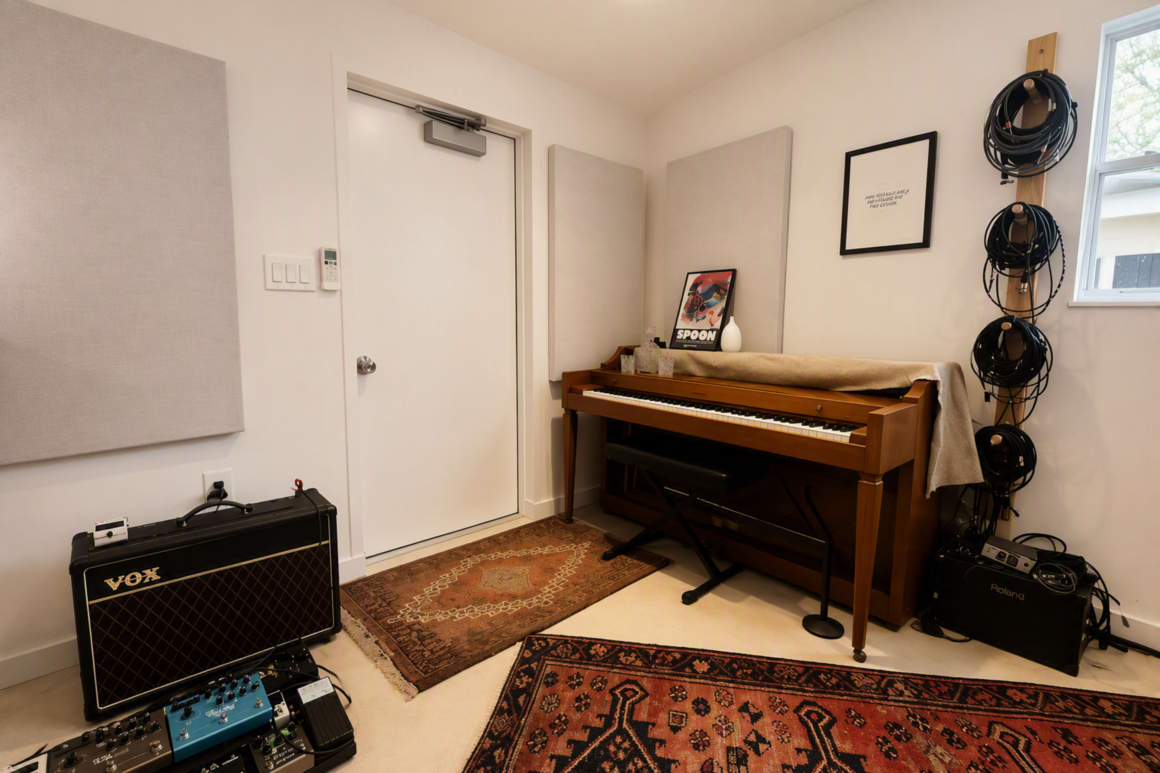 The room features a piano against the wall, surrounded by soundproofing panels, a door, and various music equipment including an amplifier and pedal effects.