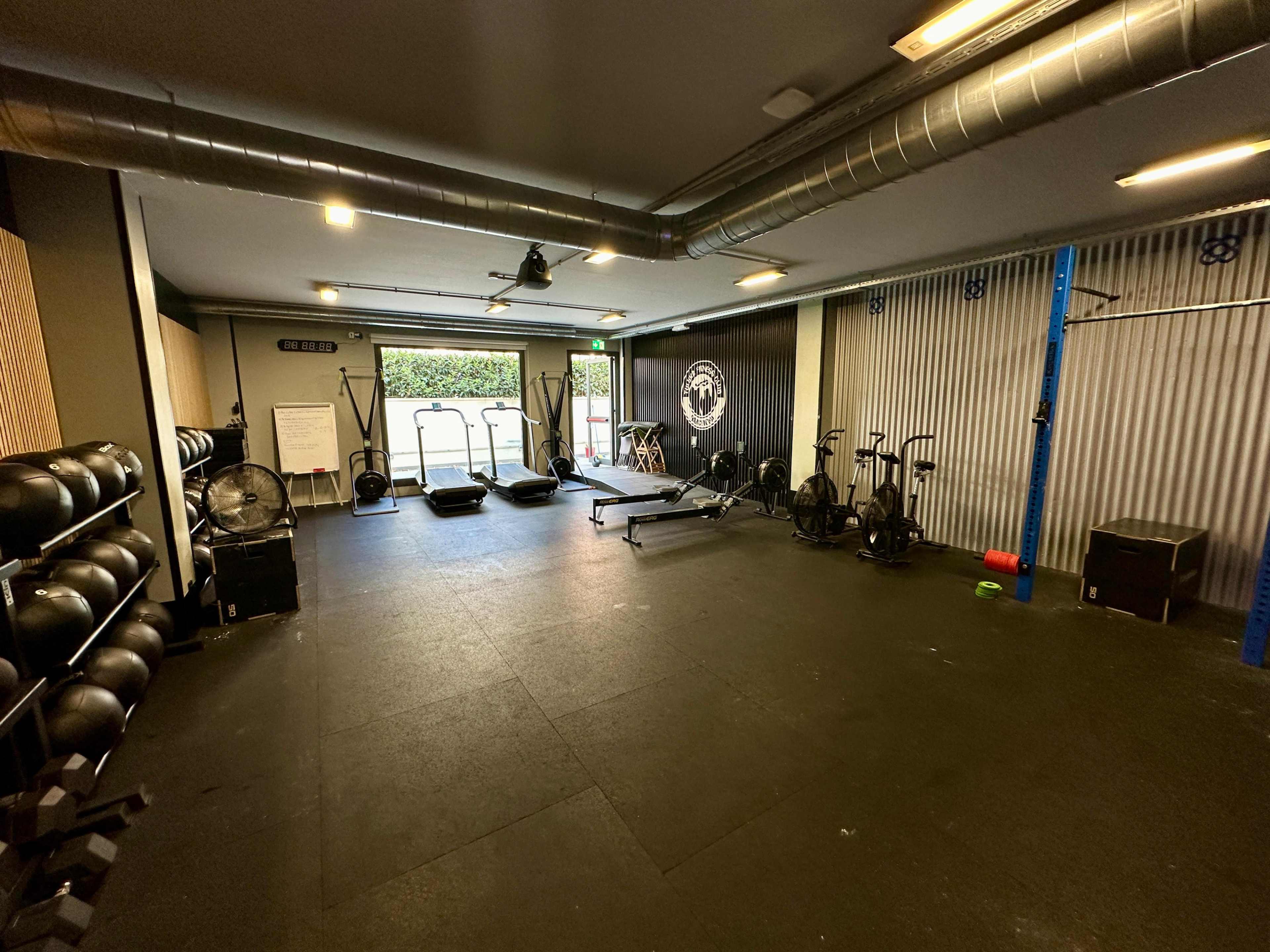 A modern gym with various exercise equipment, including weights, a rowing machine, and sleds, set in an open space with a large window.