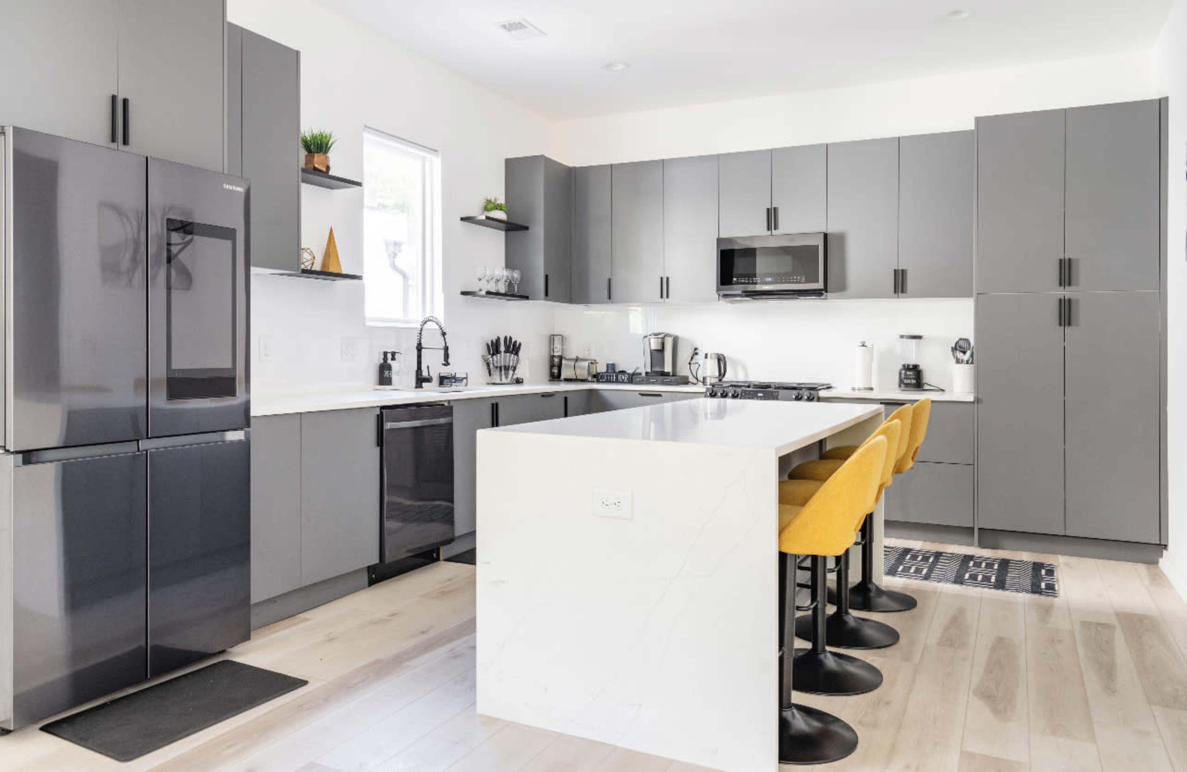 A modern kitchen features gray cabinetry, stainless steel appliances, a white island with yellow bar stools, and ample counter space.