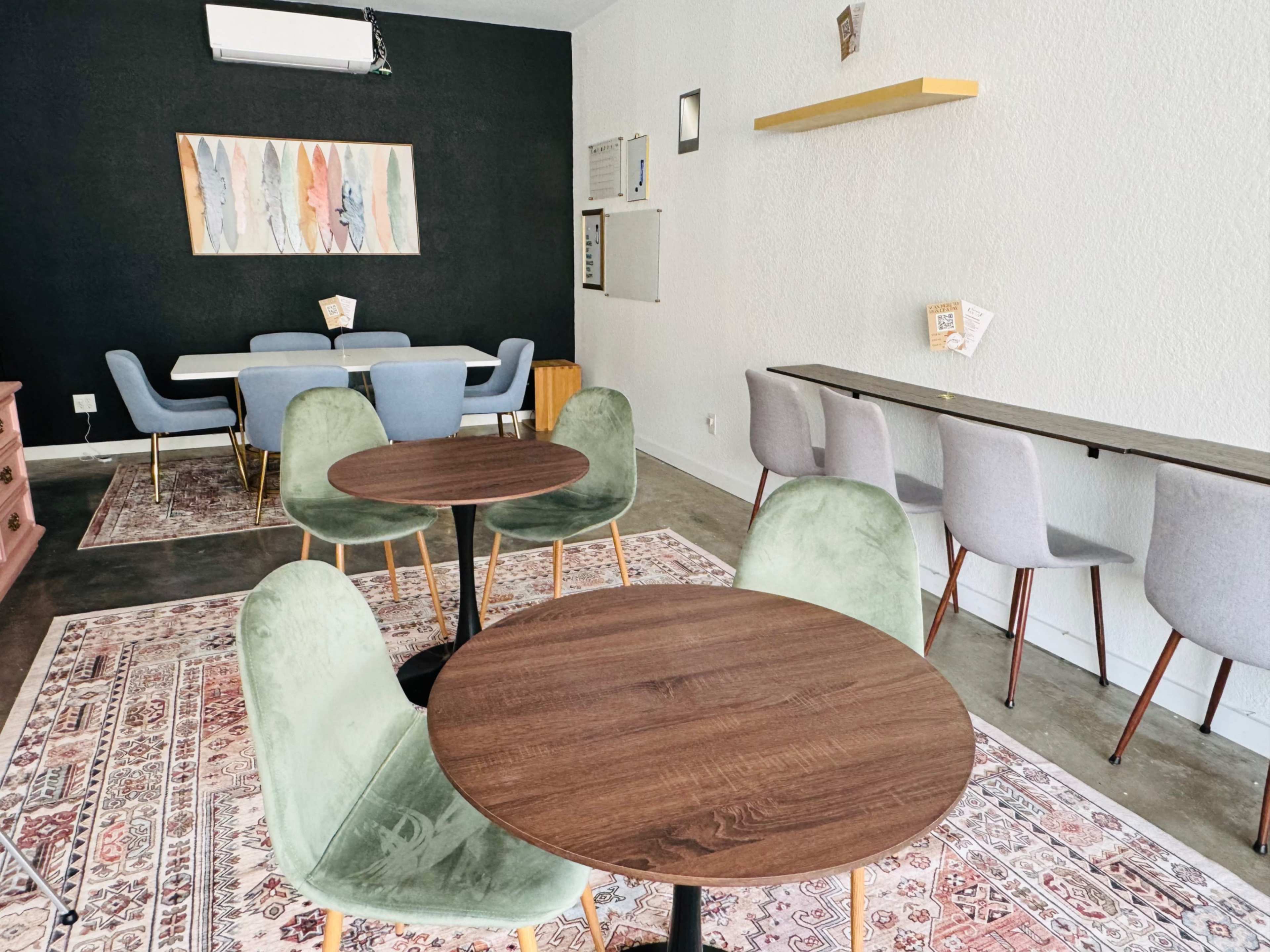 A modern café interior with wooden tables, green and gray chairs, a patterned rug, and artwork on the wall.