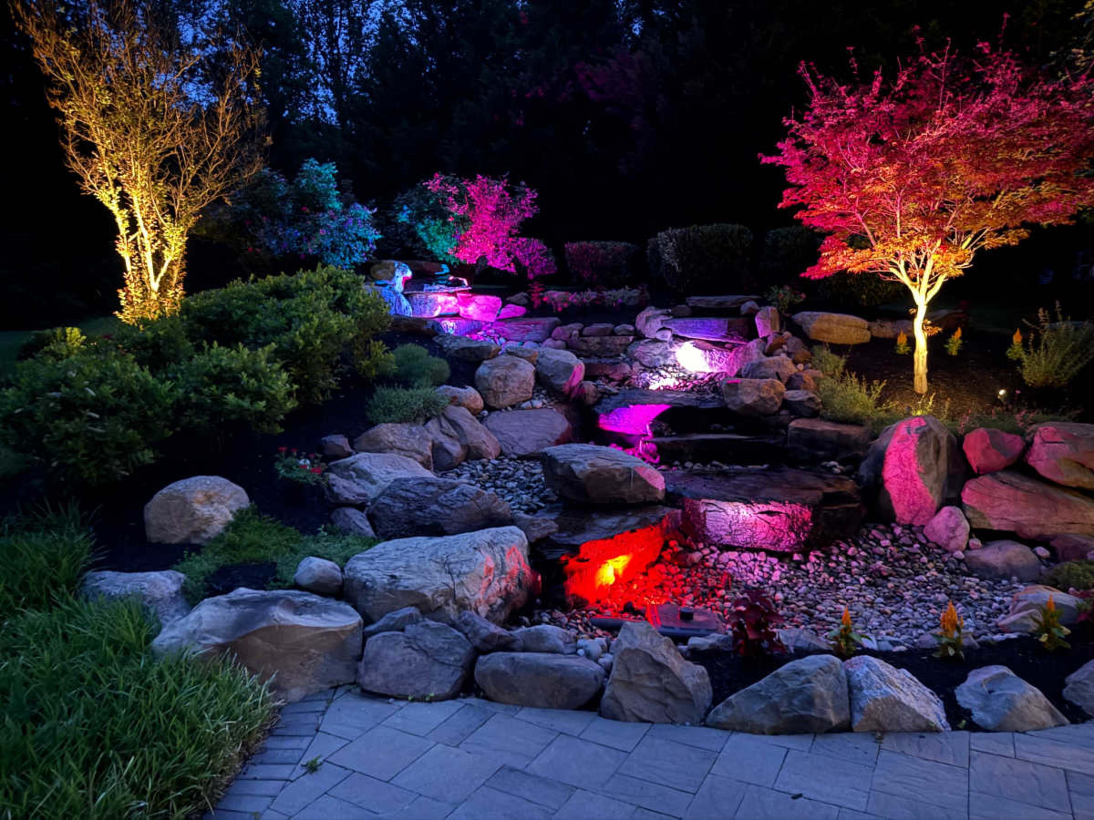 The image showcases a landscaped garden with illuminated rocks, a water feature, and colored trees set against a twilight sky.