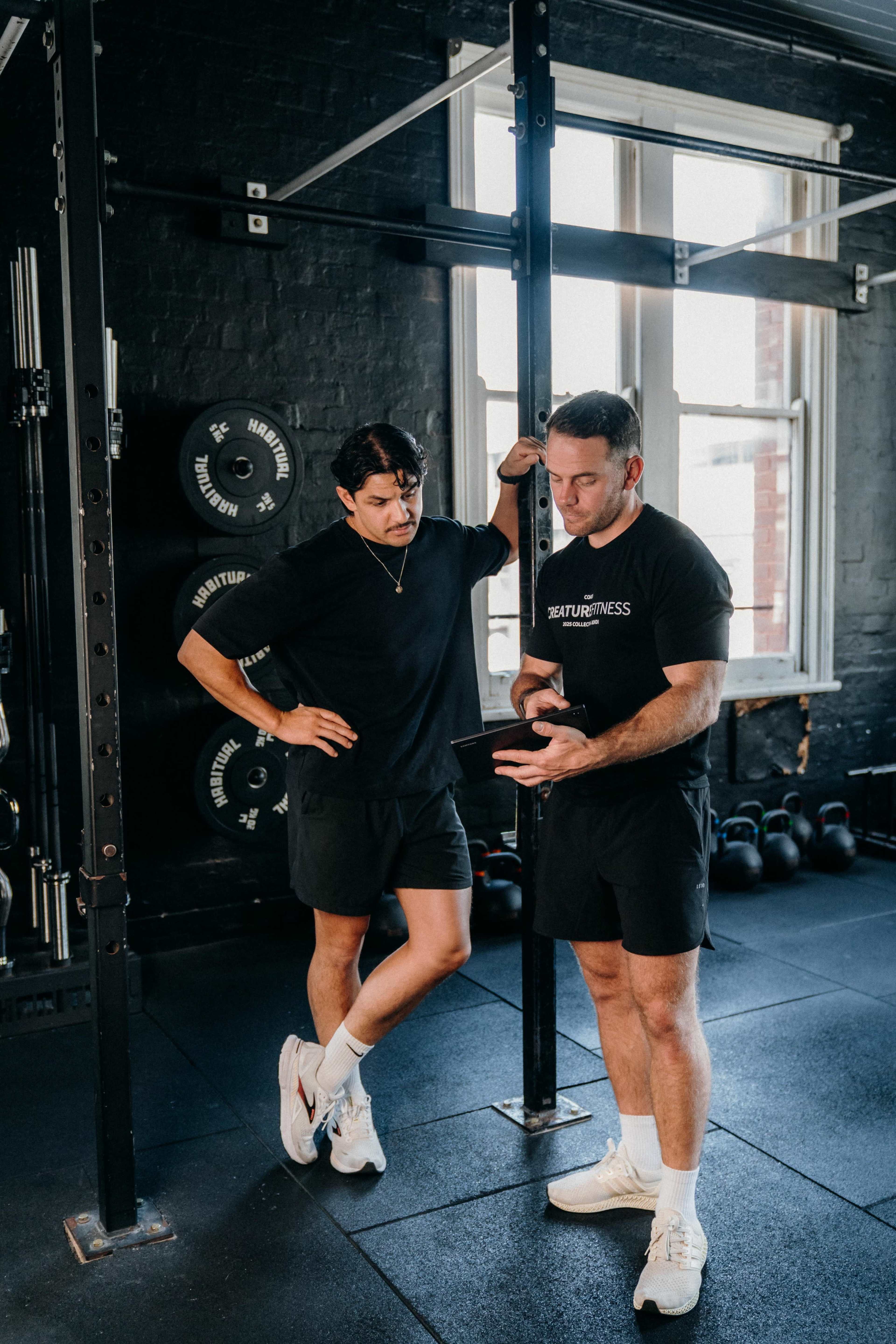 Two men are standing in a gym, one is leaning against a pillar while the other is looking at a tablet.