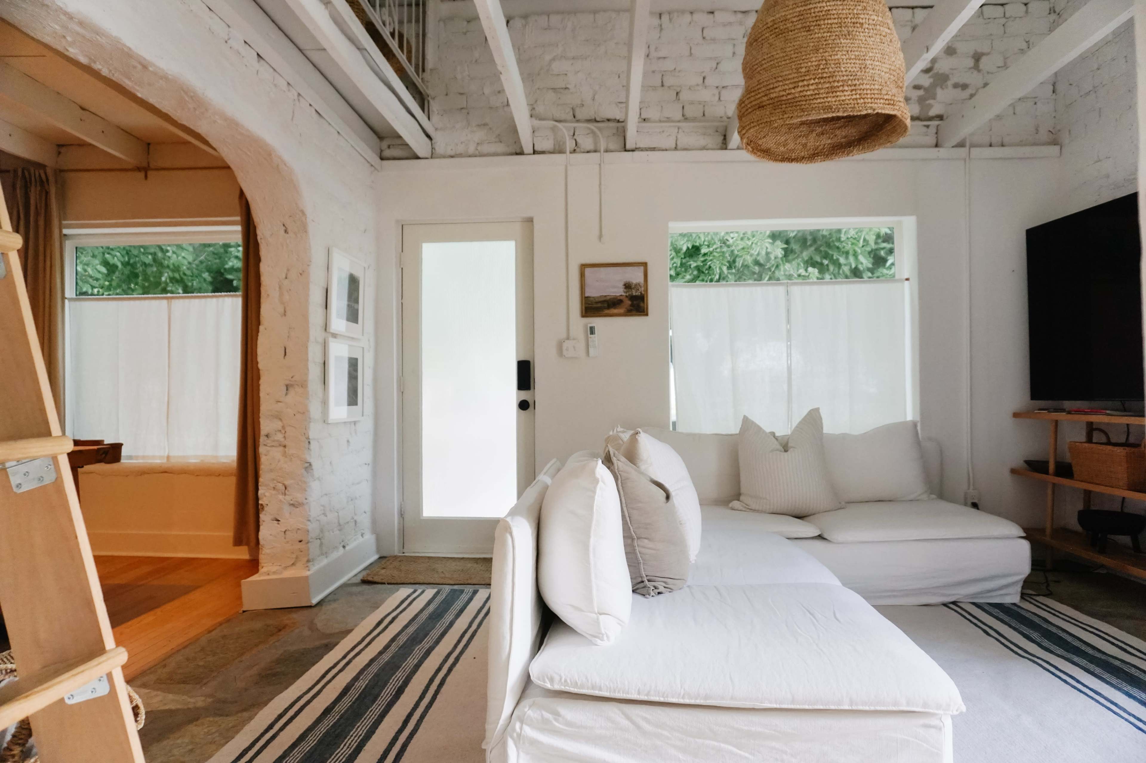 The image shows a spacious, minimalist living room with a white couch, a large window, and wooden accents.