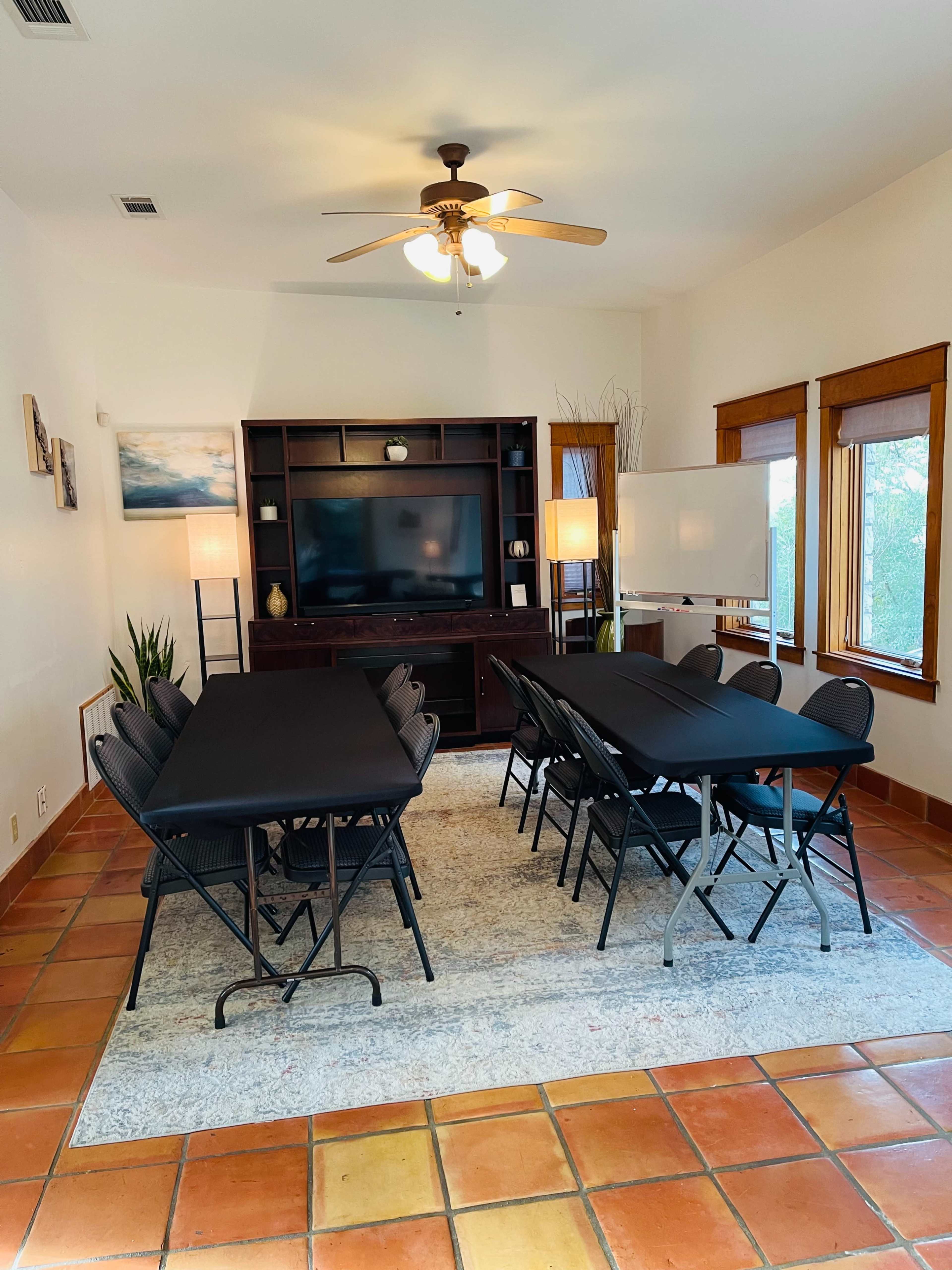 A spacious meeting room features a long arrangement of black folding tables surrounded by chairs, with a television and decorative elements in the background.