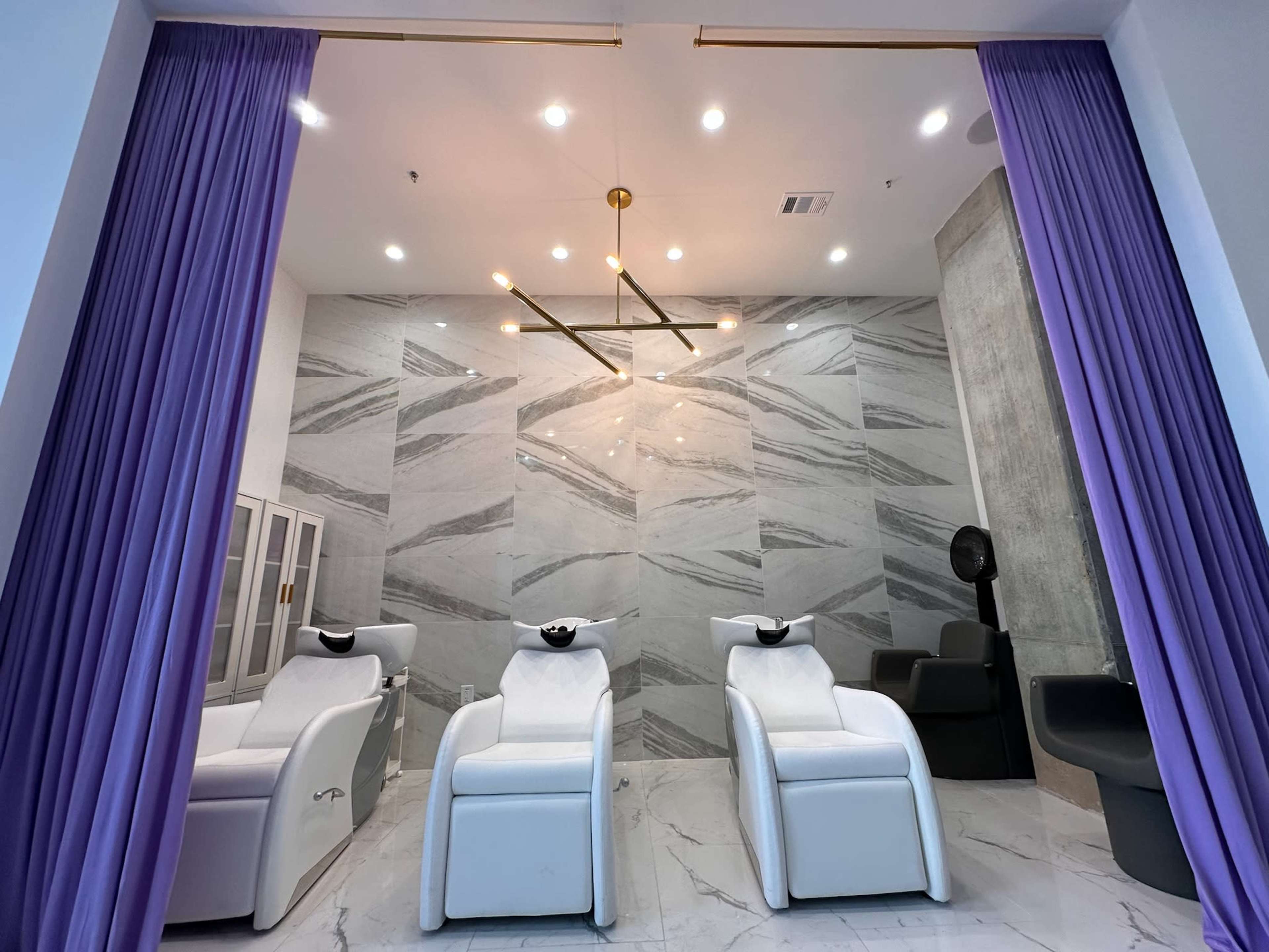 The image shows a modern hair salon interior with three white reclining chairs in front of a marble wall, separated by purple curtains.