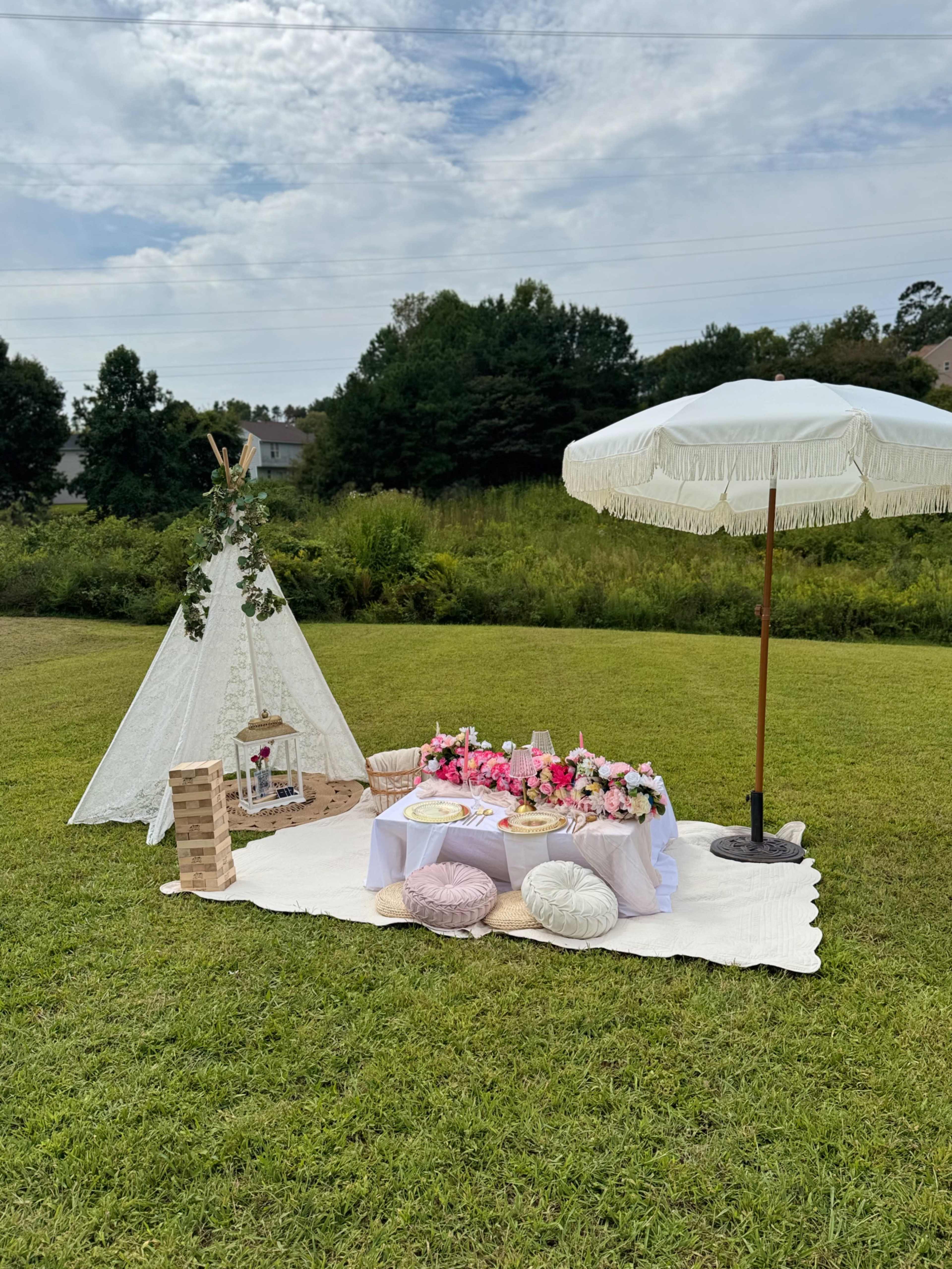 A picnic setup featuring a teepee, a decorative table with flowers, and a large umbrella on a grassy field.