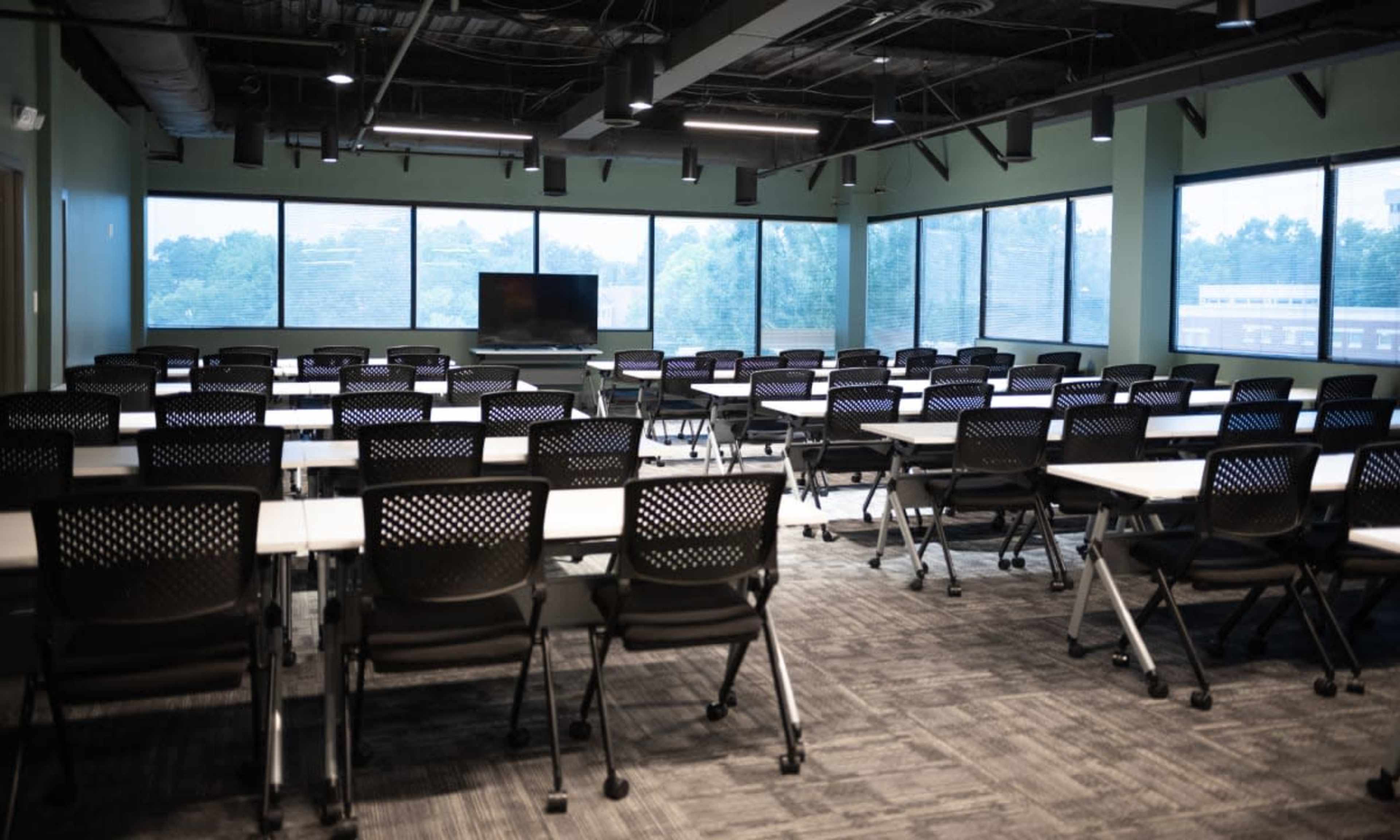 The image shows a modern conference room with rows of chairs and tables arranged for a meeting, large windows providing natural light and views of the outside.