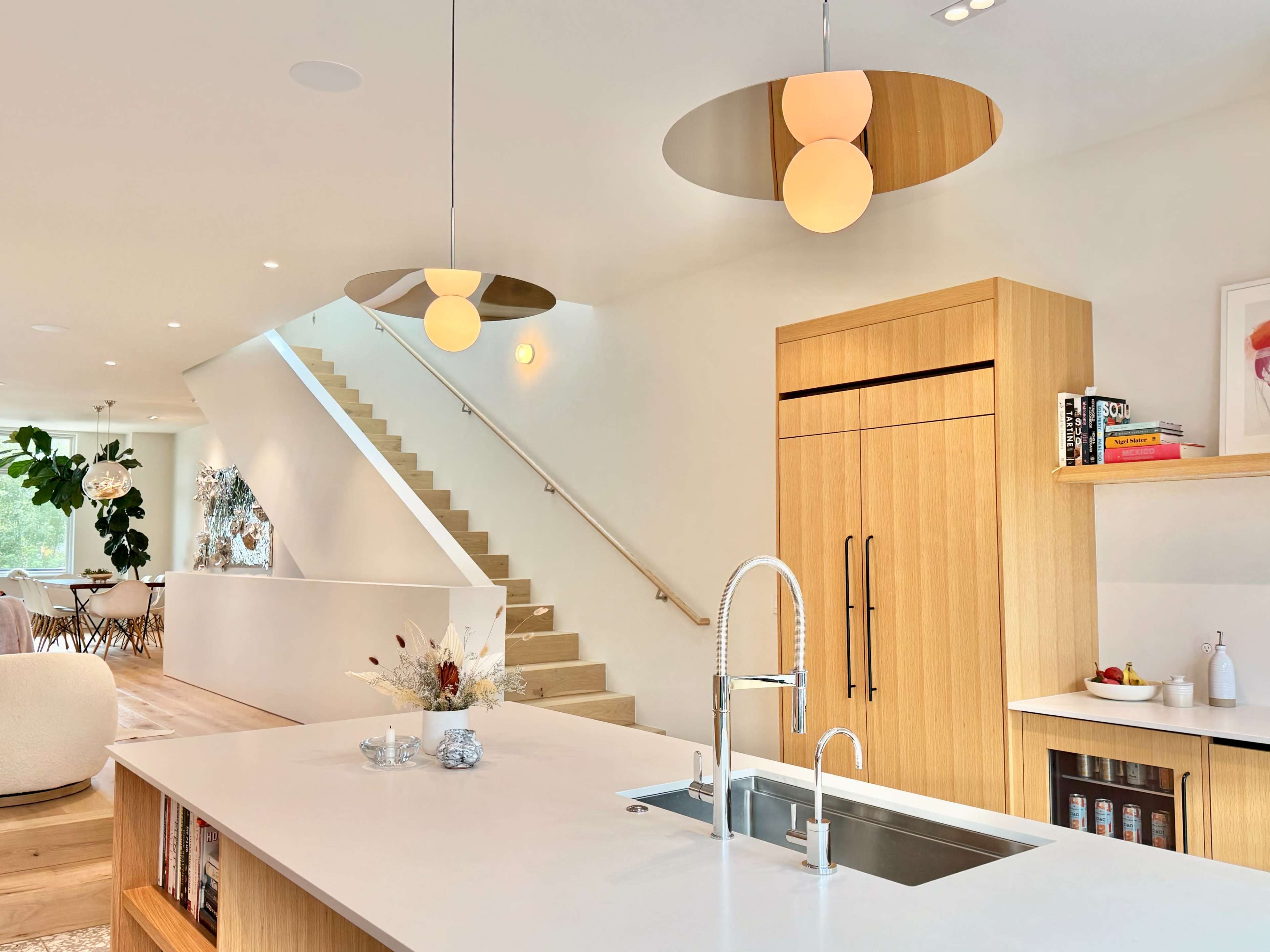 The image shows a modern kitchen with a wooden island, a large sink, and minimalist pendant lighting, alongside a staircase leading to an upper level.