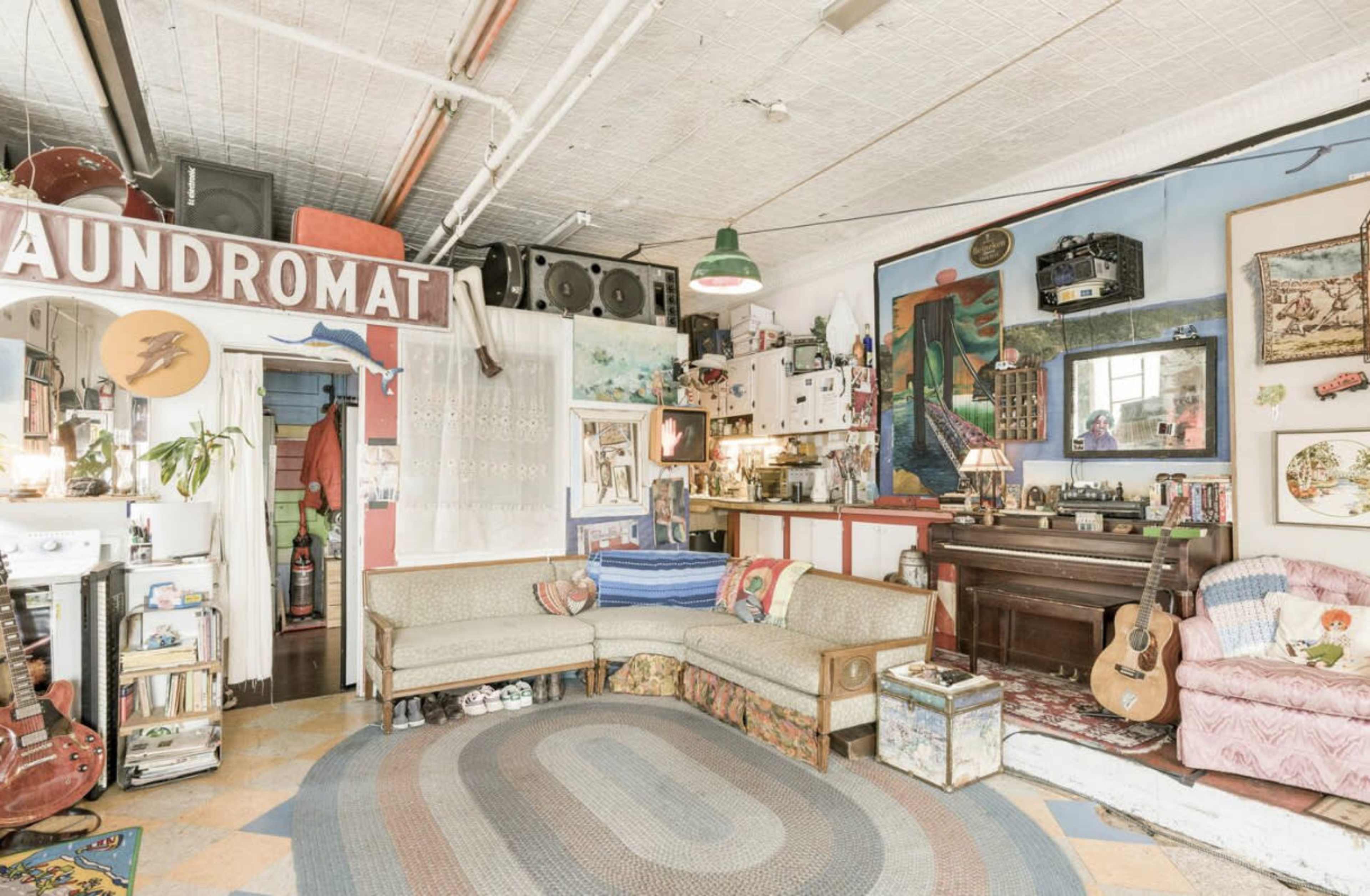 A brightly lit, eclectic living space featuring a cozy couch, a vintage piano, and various artworks and decorations on the walls.