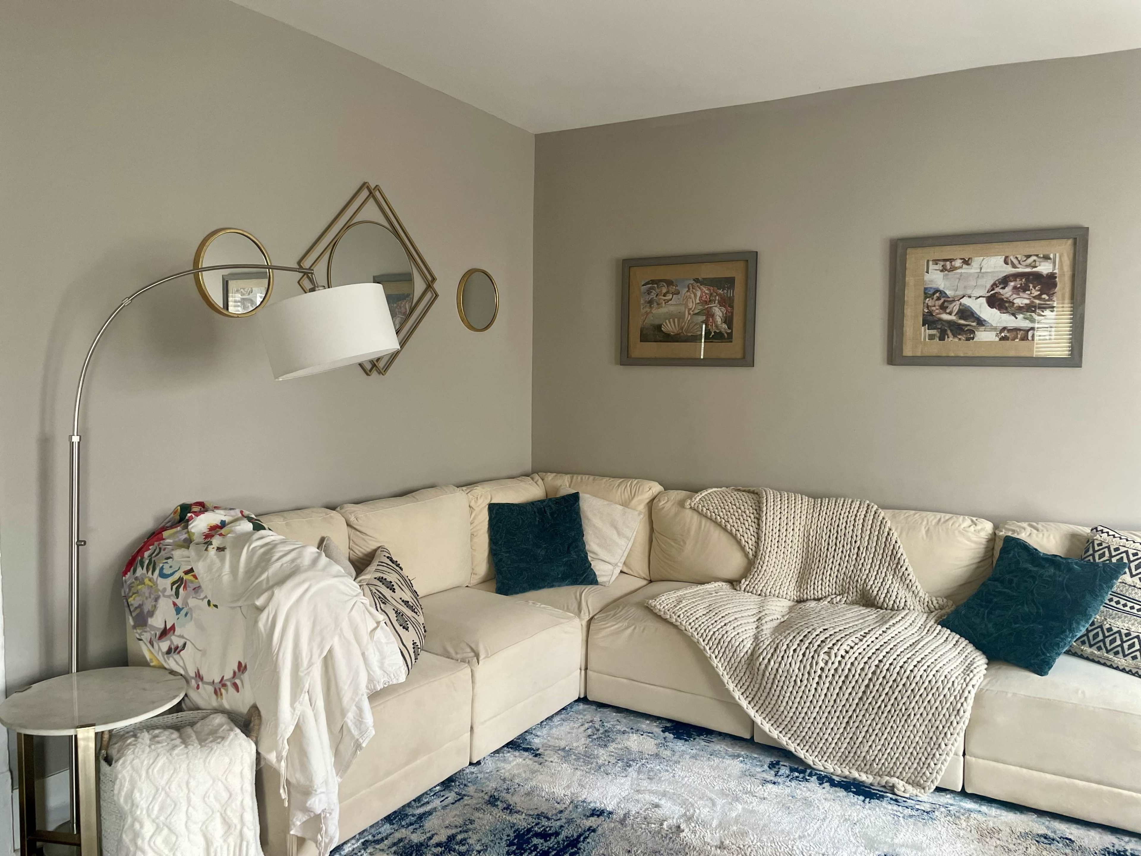 The image shows a cozy living room with a sectional couch, a floor lamp, decorative mirrors, and framed artwork on the walls.