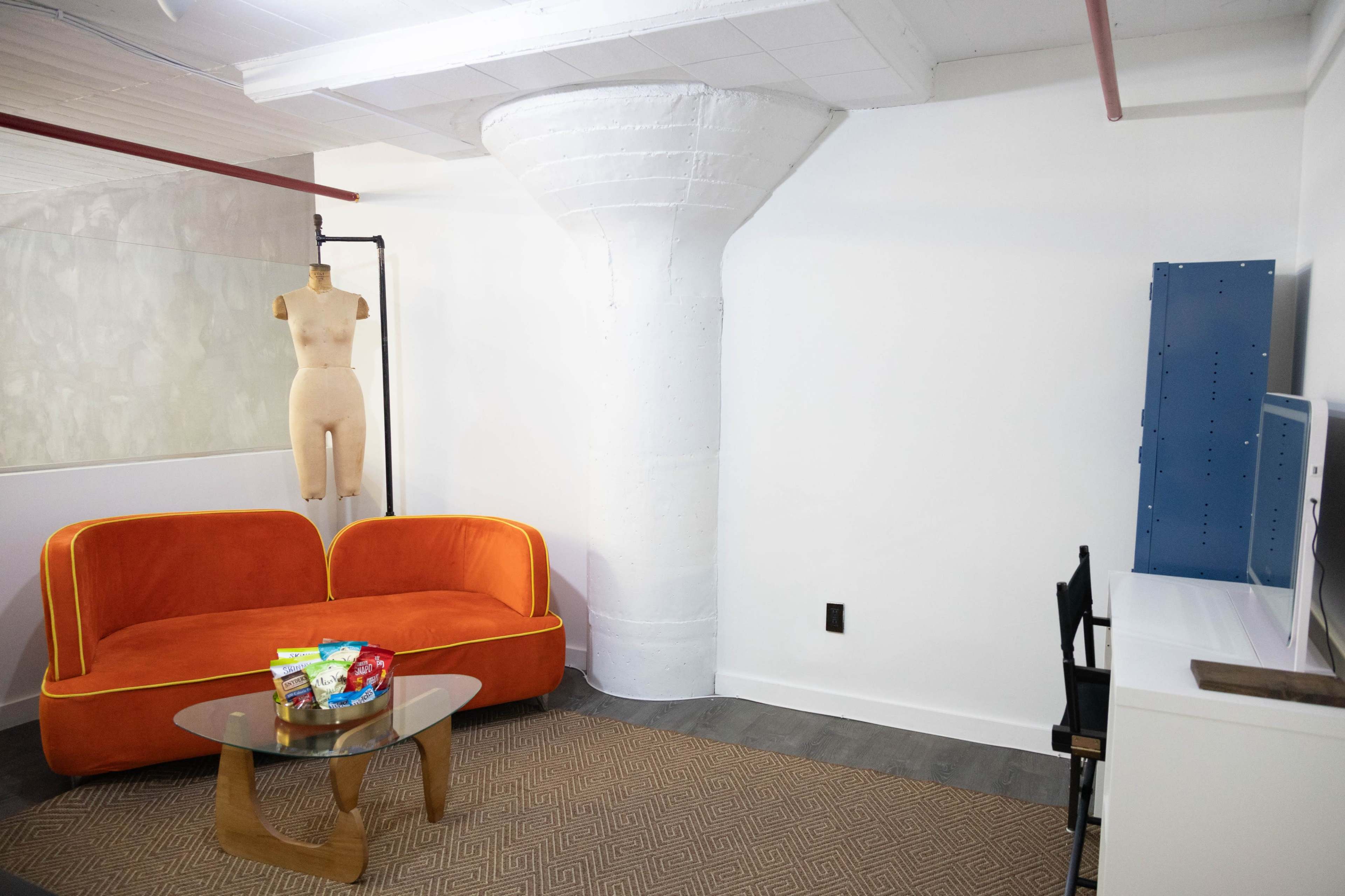 The image shows a small room featuring an orange couch, a glass coffee table with snacks, a mannequin, and a white column.