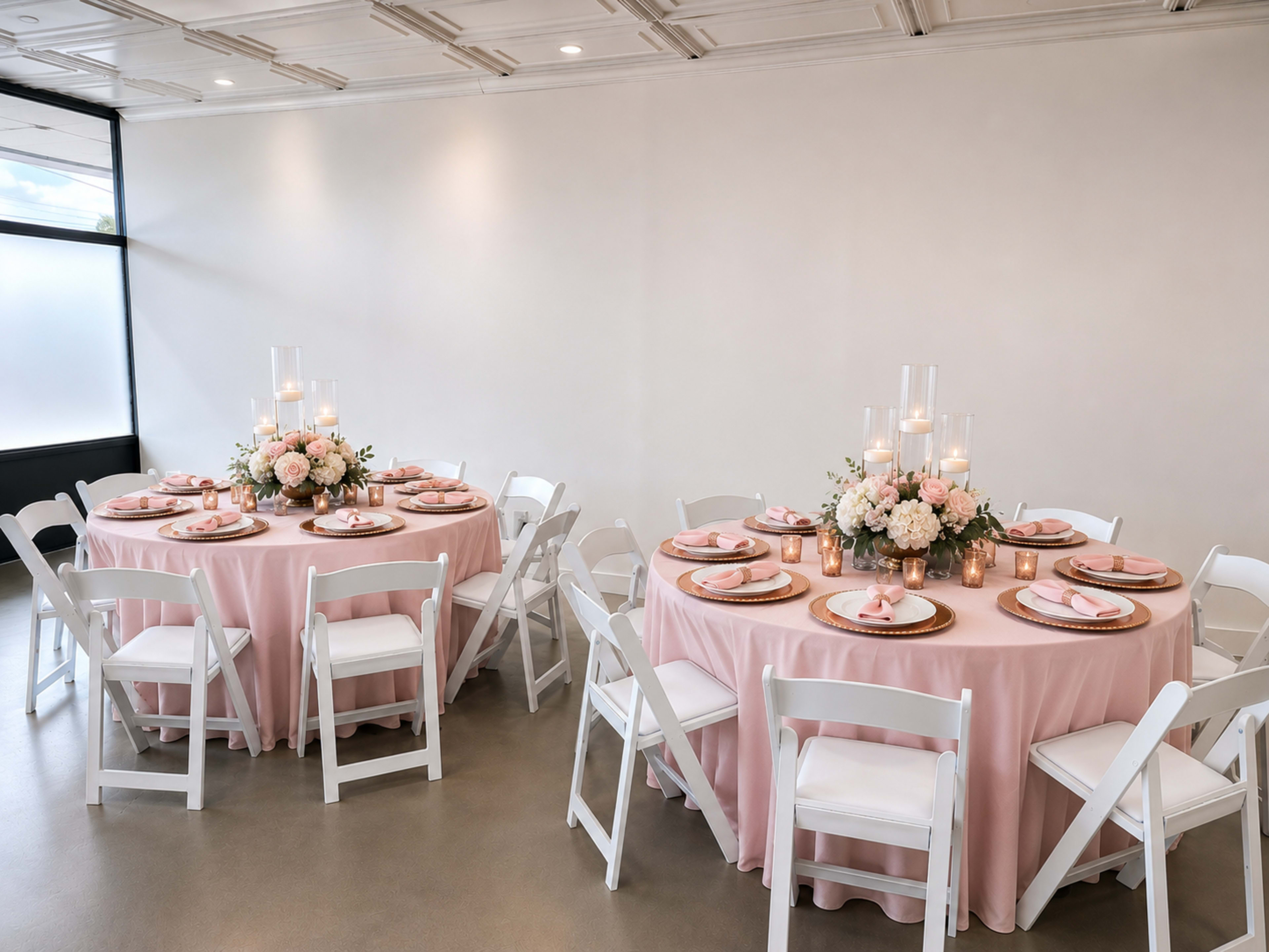 Two round tables draped in pink tablecloths are set with floral centerpieces and lit candles, surrounded by white chairs.