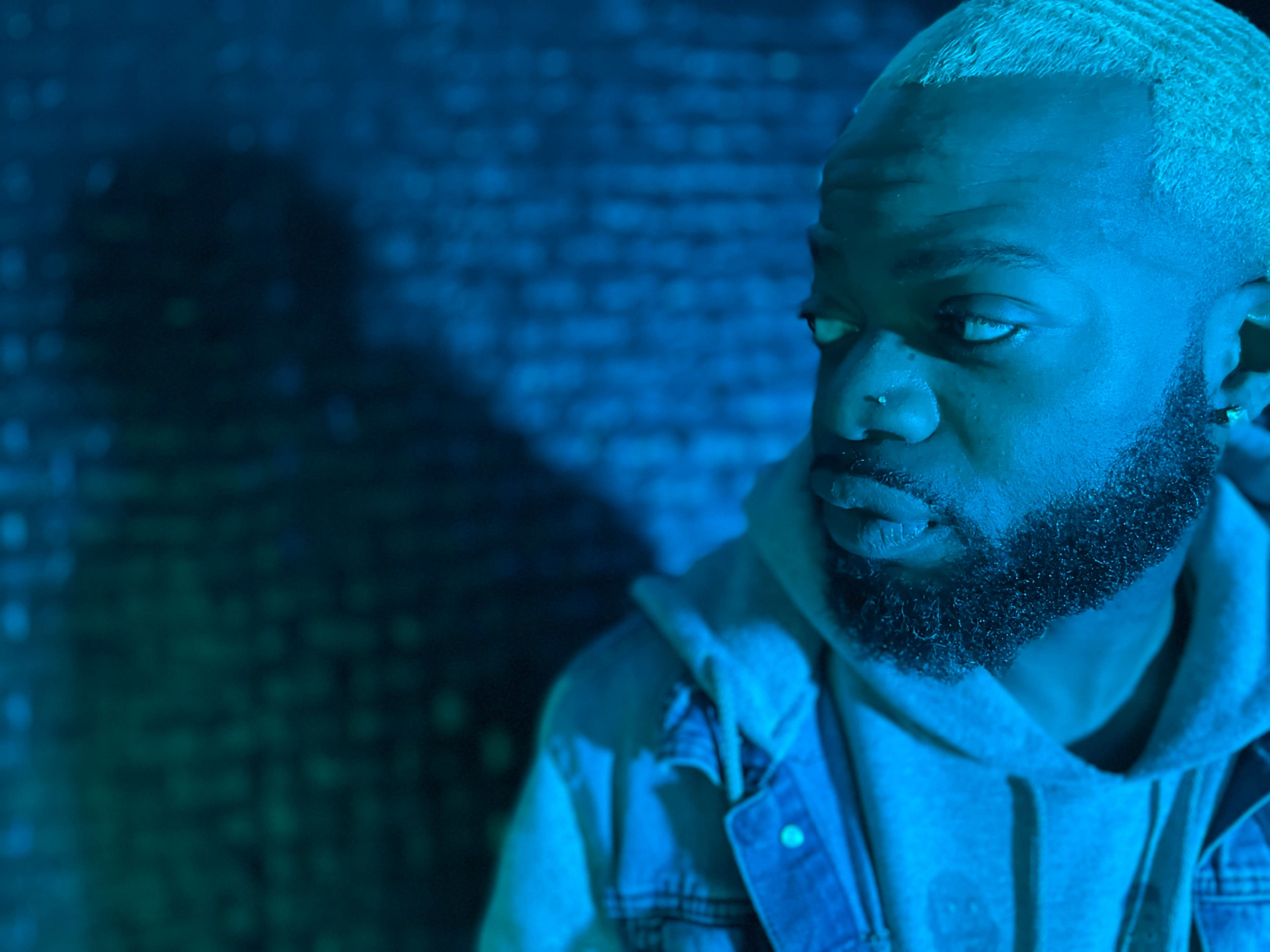A person with light-colored hair and a beard gazes to the side against a dark, textured background illuminated by blue light.