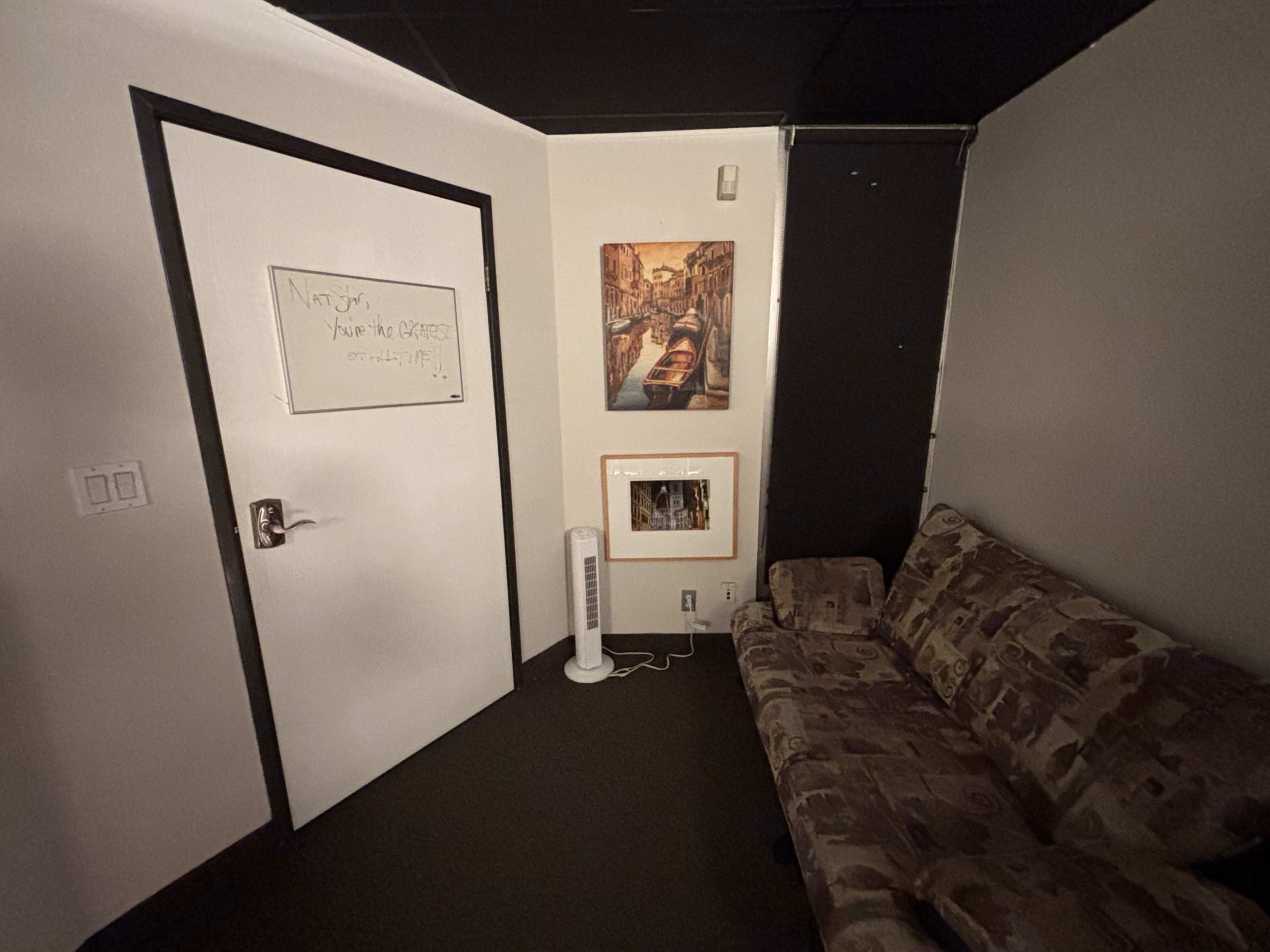 The image shows a small room with a couch, a door with a whiteboard, and a framed picture on the wall beside a floor fan.