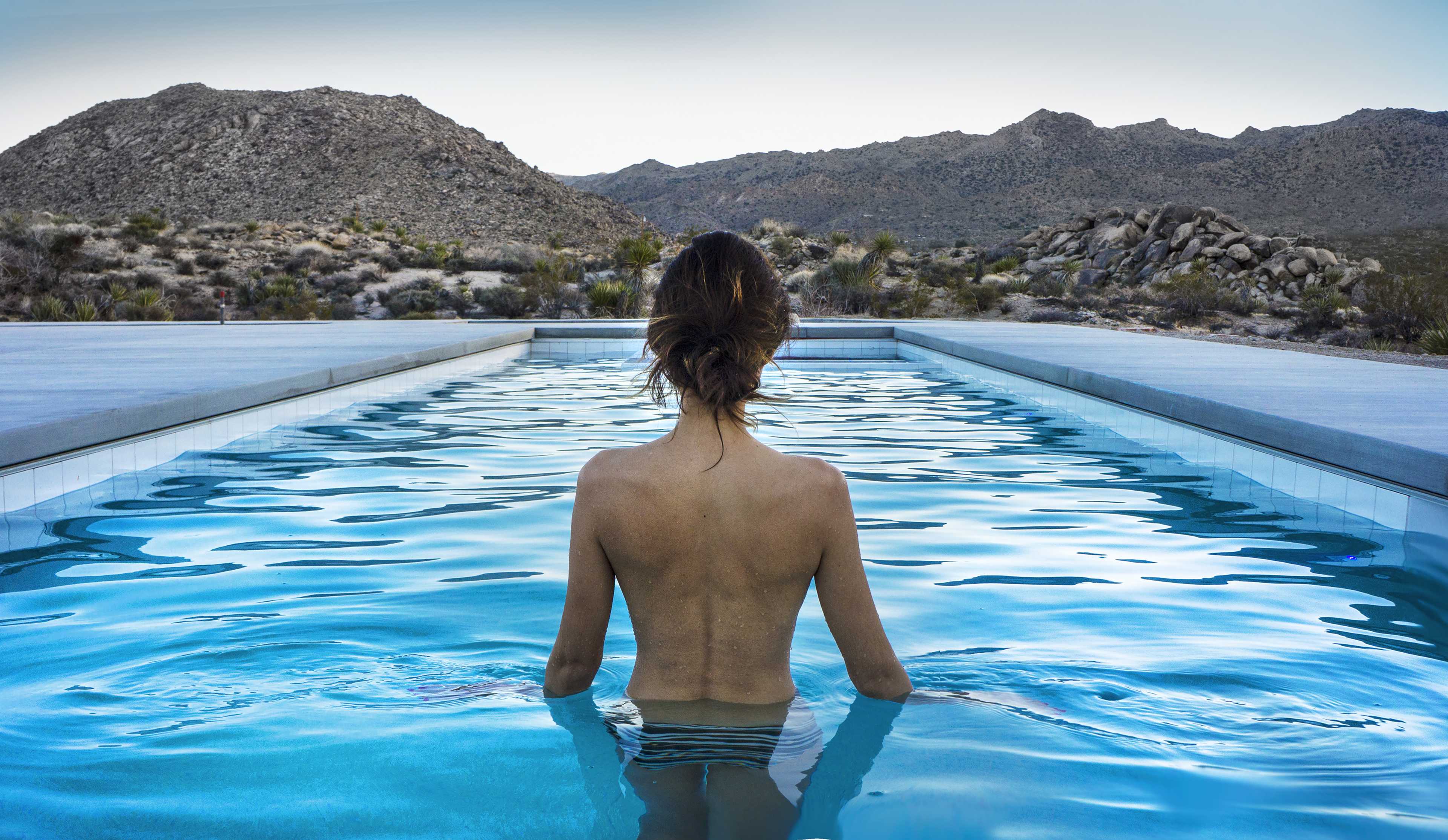 A person stands in a swimming pool, facing away, with a mountainous desert landscape in the background.