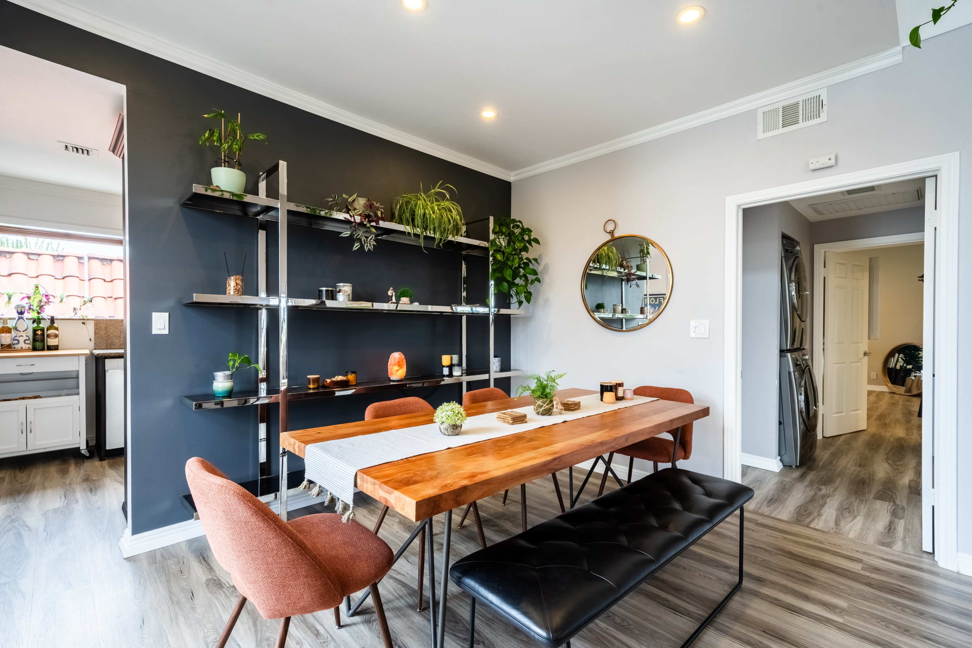 A dining area features a wooden table surrounded by upholstered chairs, facing a wall with open shelving displaying plants and decorative items.