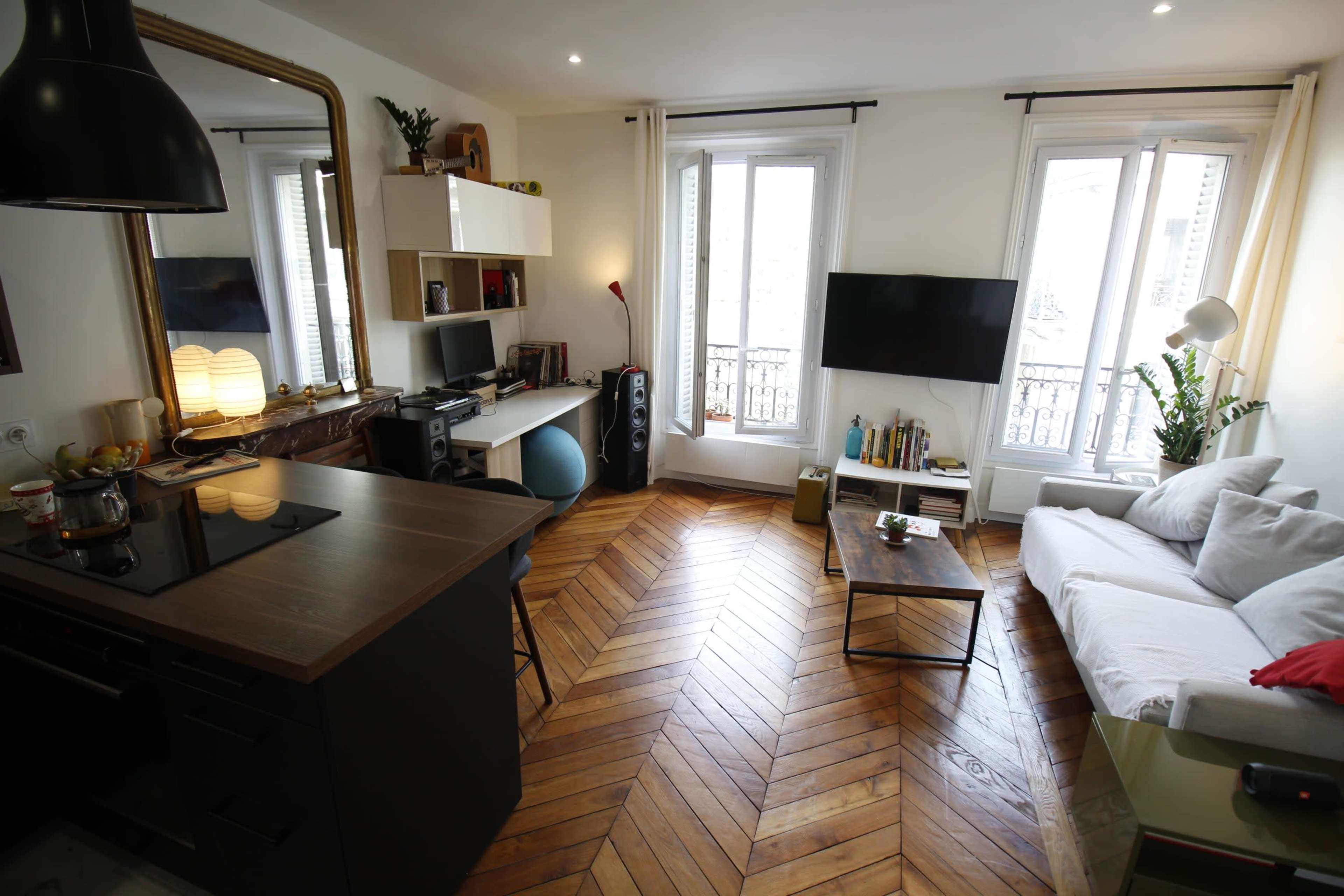 The image shows a cozy apartment featuring a kitchen area with a dark countertop, a living room with a white sofa, a coffee table, and large windows allowing natural light in.