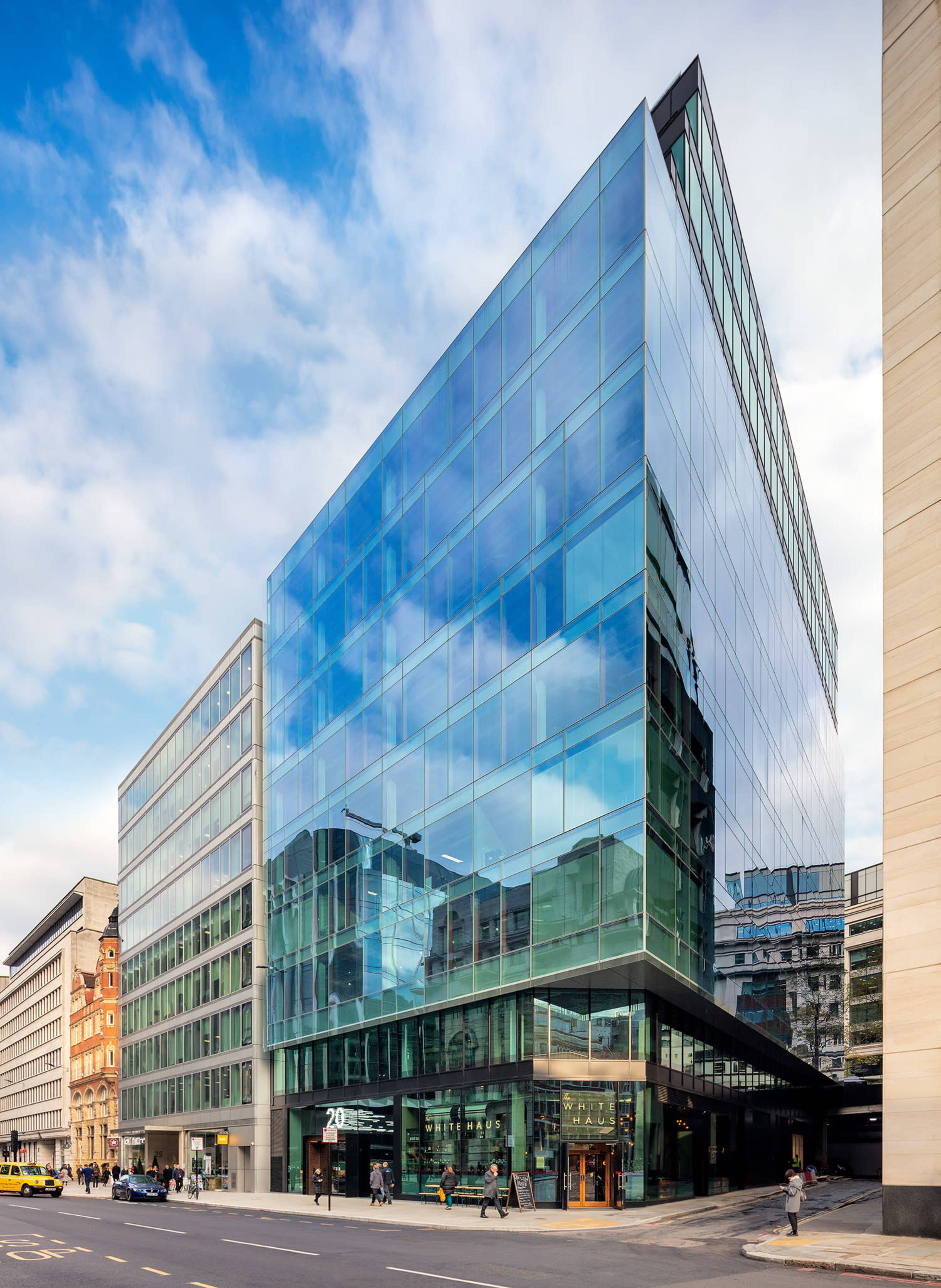 A modern glass office building with a sharp angular design stands at the intersection of two streets, reflecting the surrounding urban landscape.