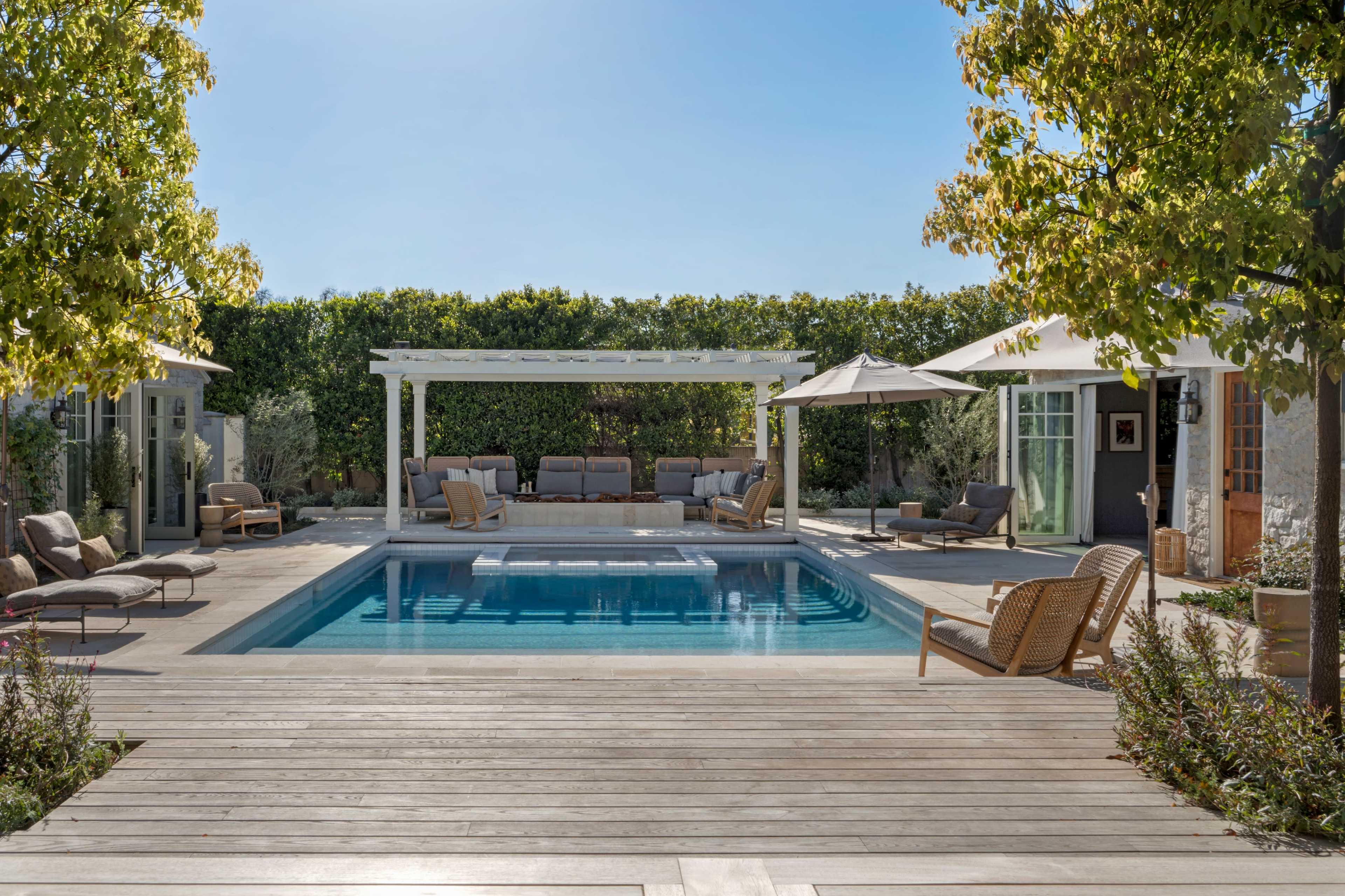 A backyard features a rectangular swimming pool surrounded by seating areas, umbrellas, and lush greenery.