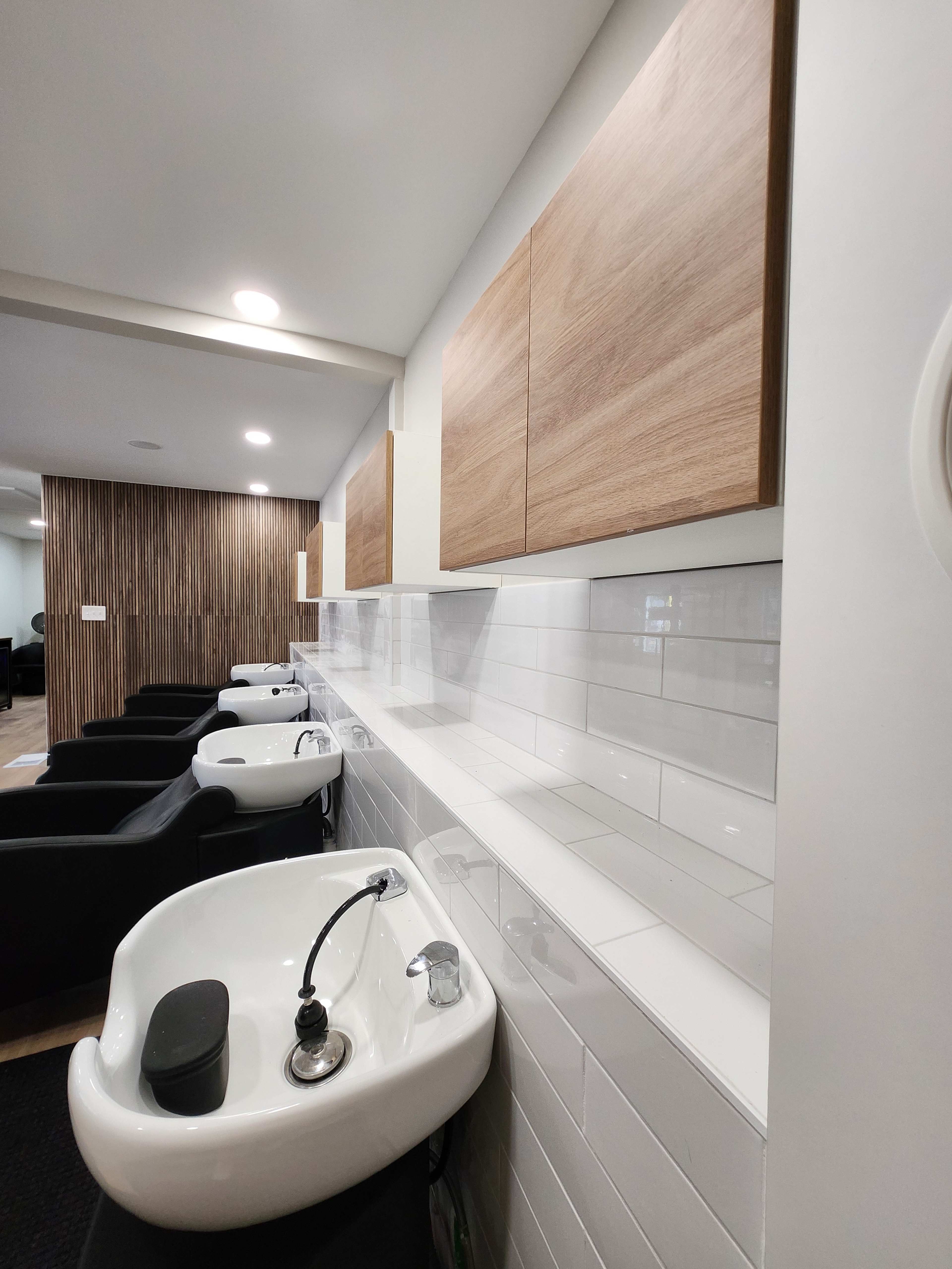 The image shows a modern salon space featuring several black shampoo bowls lined against a white and wood-paneled wall.