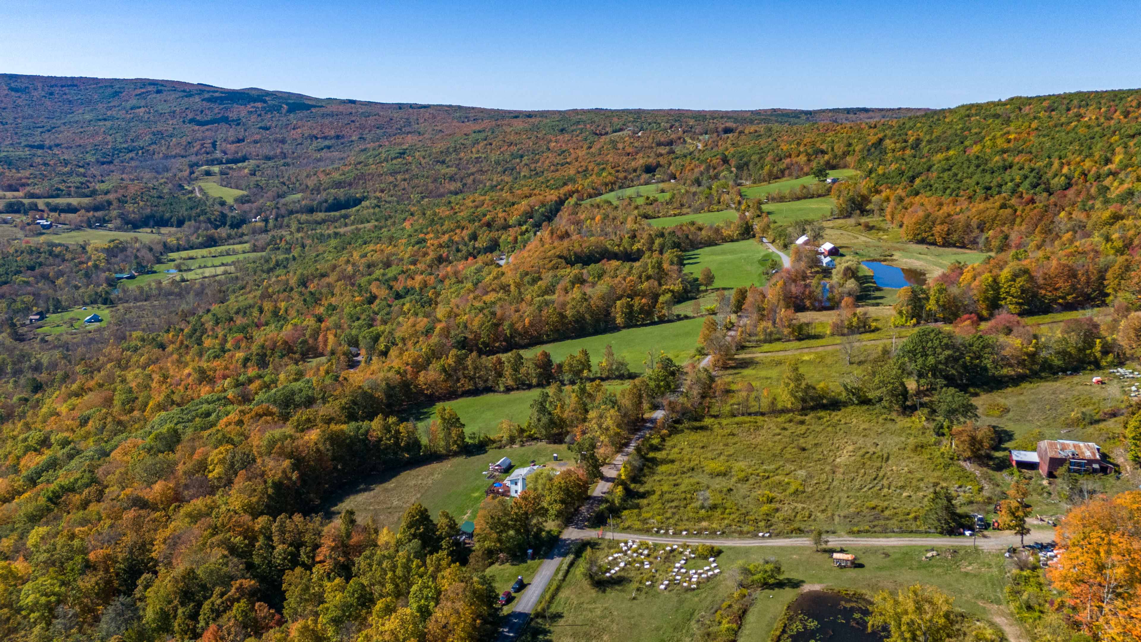The image shows a panoramic view of rolling hills and fields with autumn-colored trees, a pond, and a farmstead in a rural landscape.