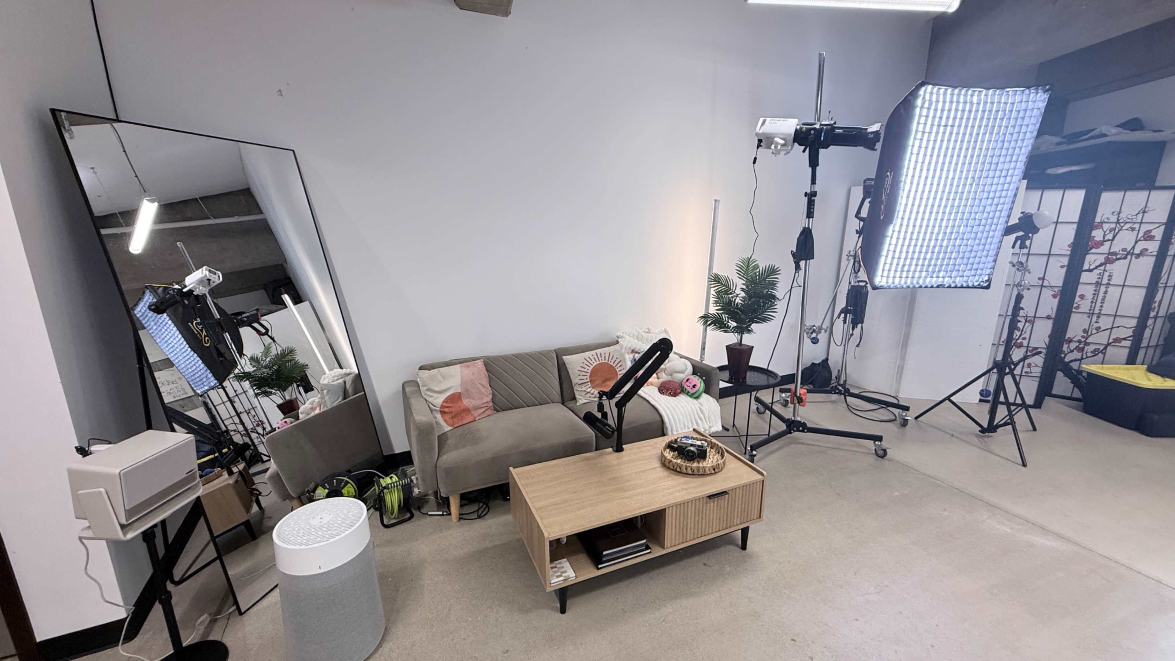 The image shows a well-lit studio space featuring a couch, a coffee table, a large mirror, and various lighting equipment arranged against a plain wall.