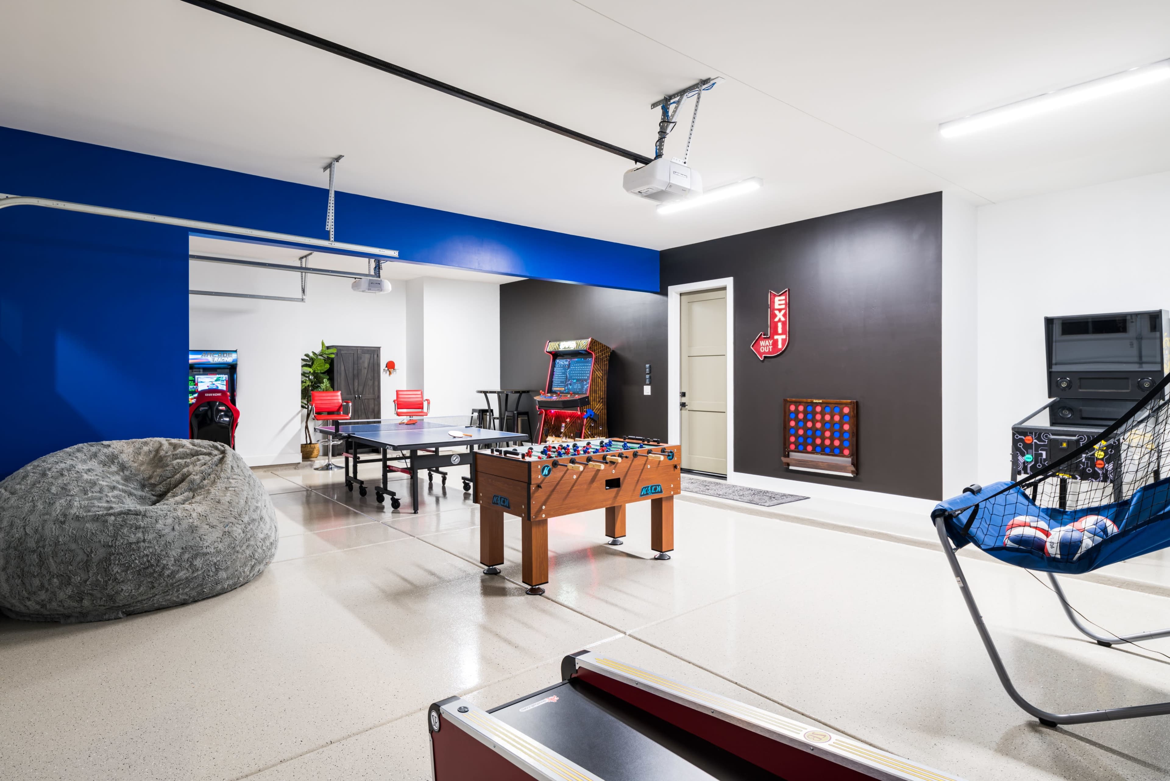 The image shows a game room featuring a foosball table, arcade games, a bean bag chair, and a hammock in a spacious, brightly lit space with blue and dark walls.