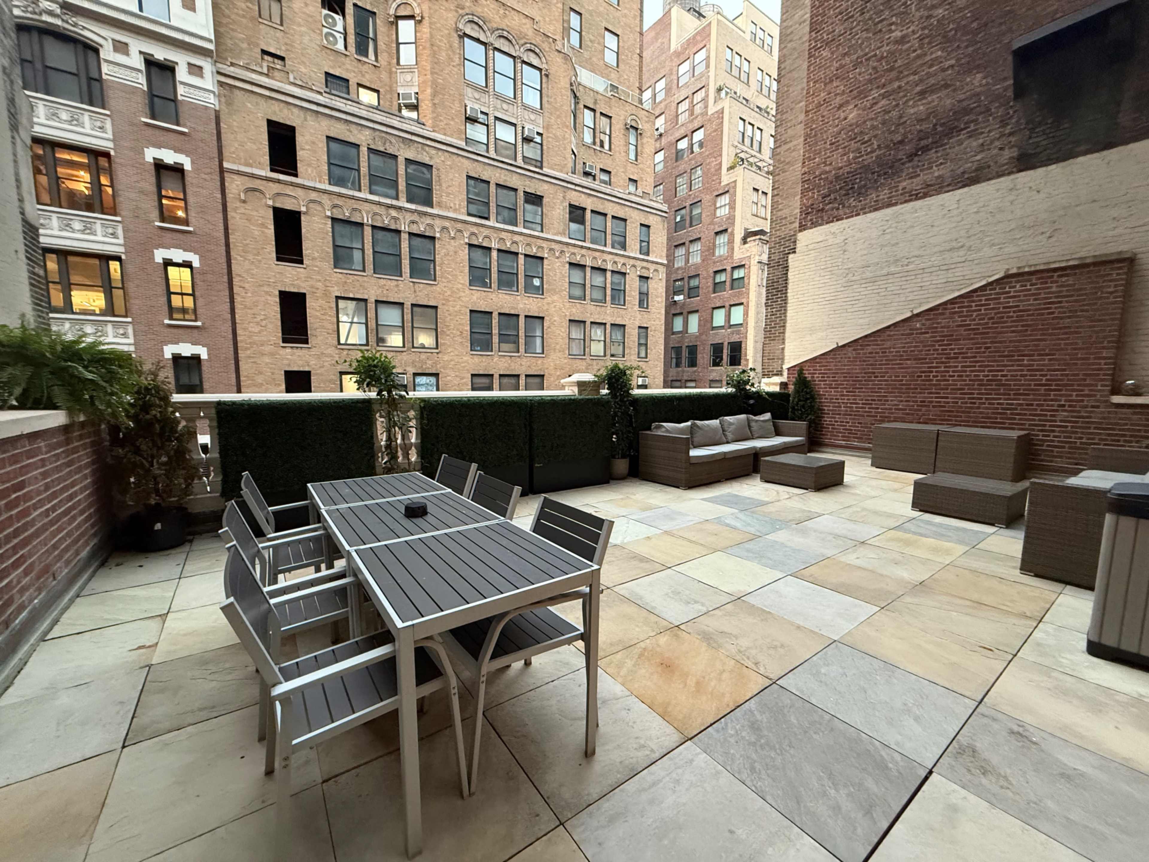 The image shows a modern rooftop terrace with a seating area, a dining table, and planters, surrounded by tall brick buildings.