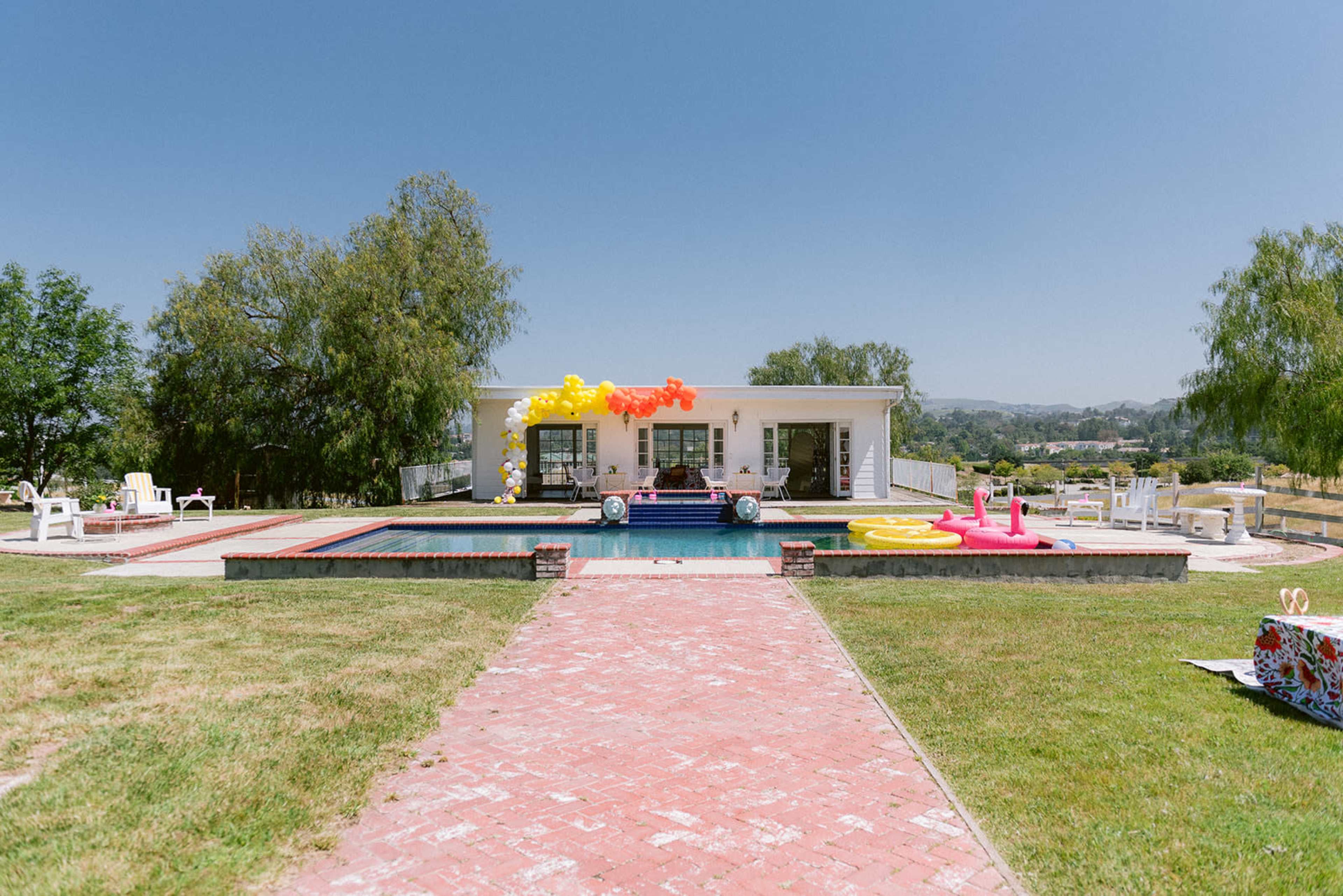 A rectangular pool surrounded by a lawn and a brick pathway leads to a white building adorned with colorful decorations.