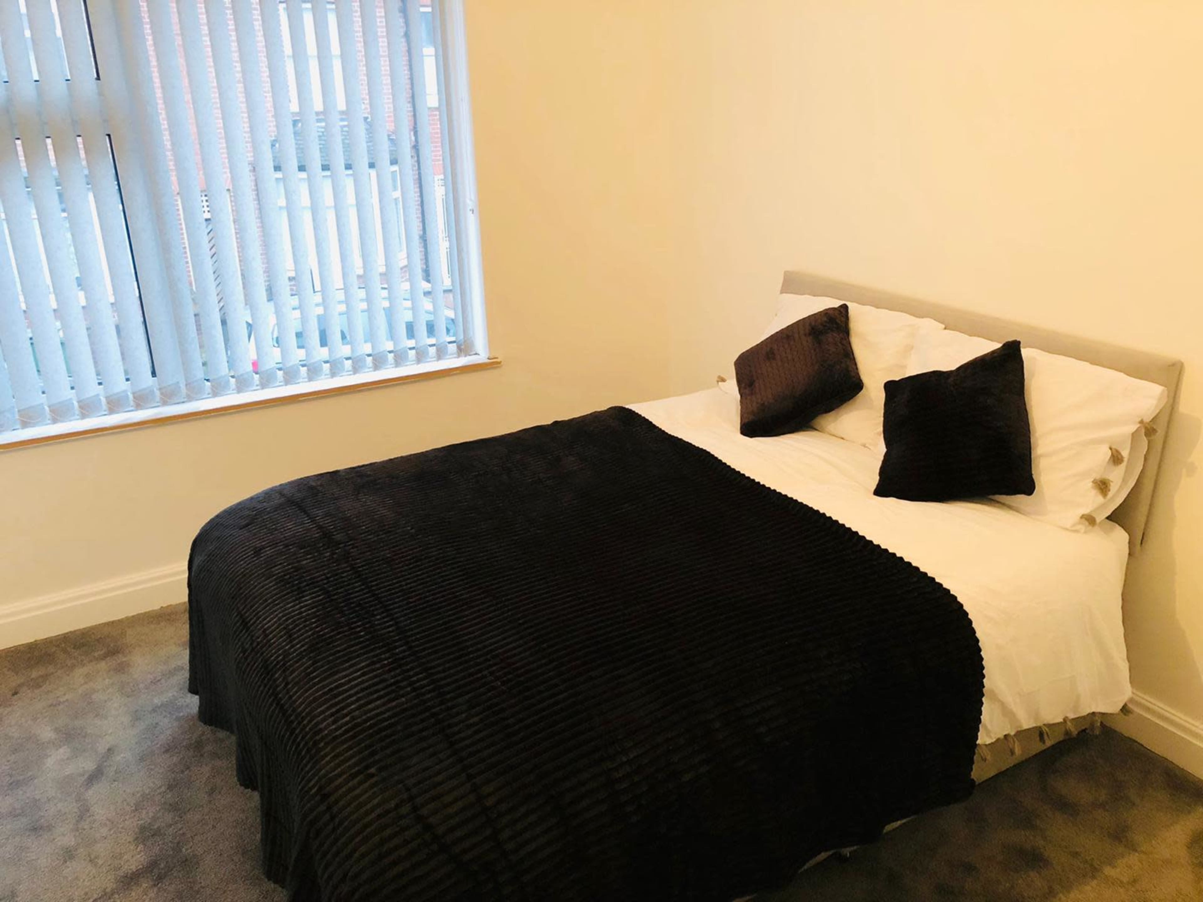 A neatly made bed with a black blanket and two black pillows sits in a room with vertical blinds and gray carpet.