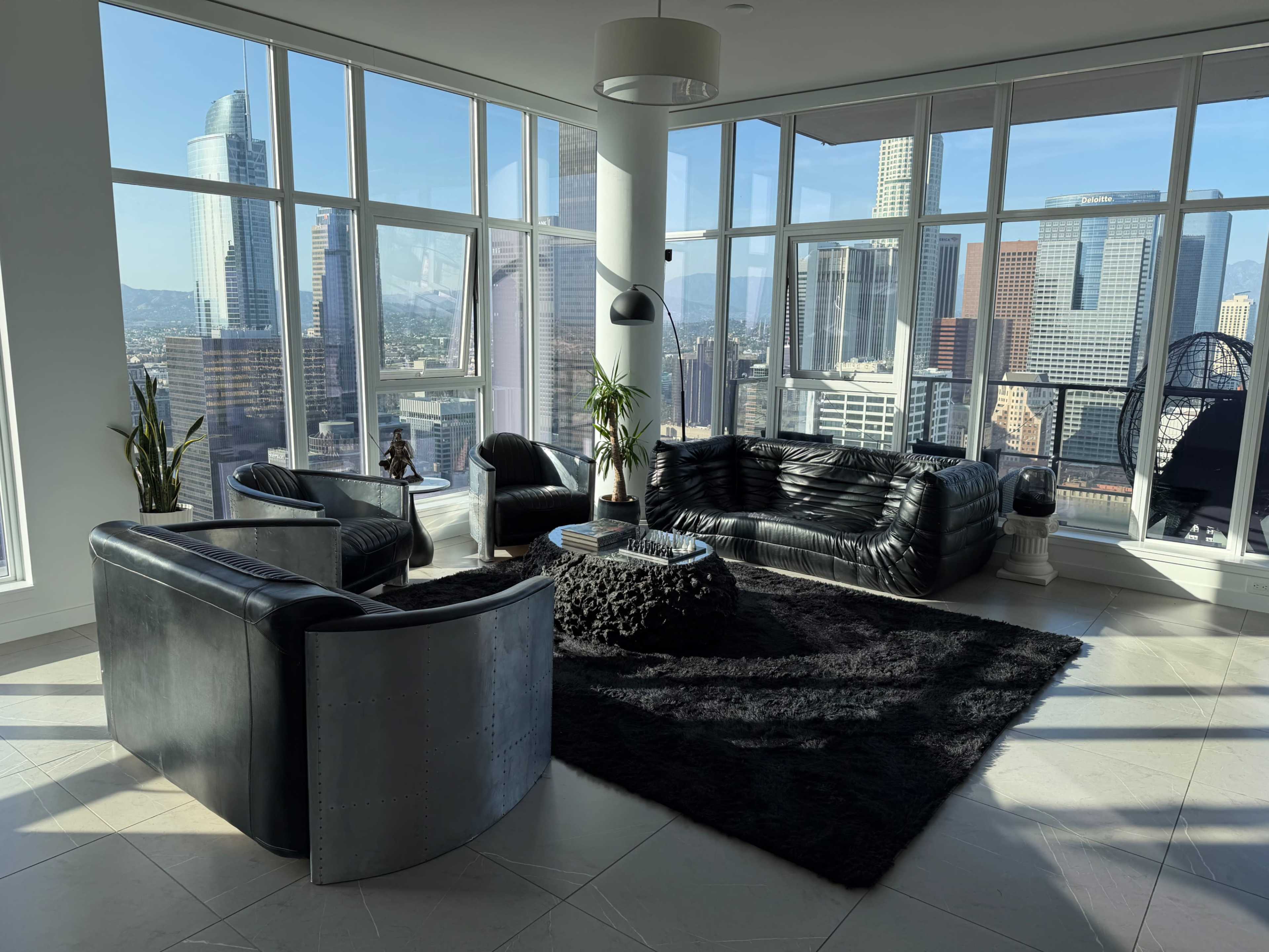 The image shows a modern living room with black furniture, large windows revealing a city skyline, and a circular rug on a light tile floor.