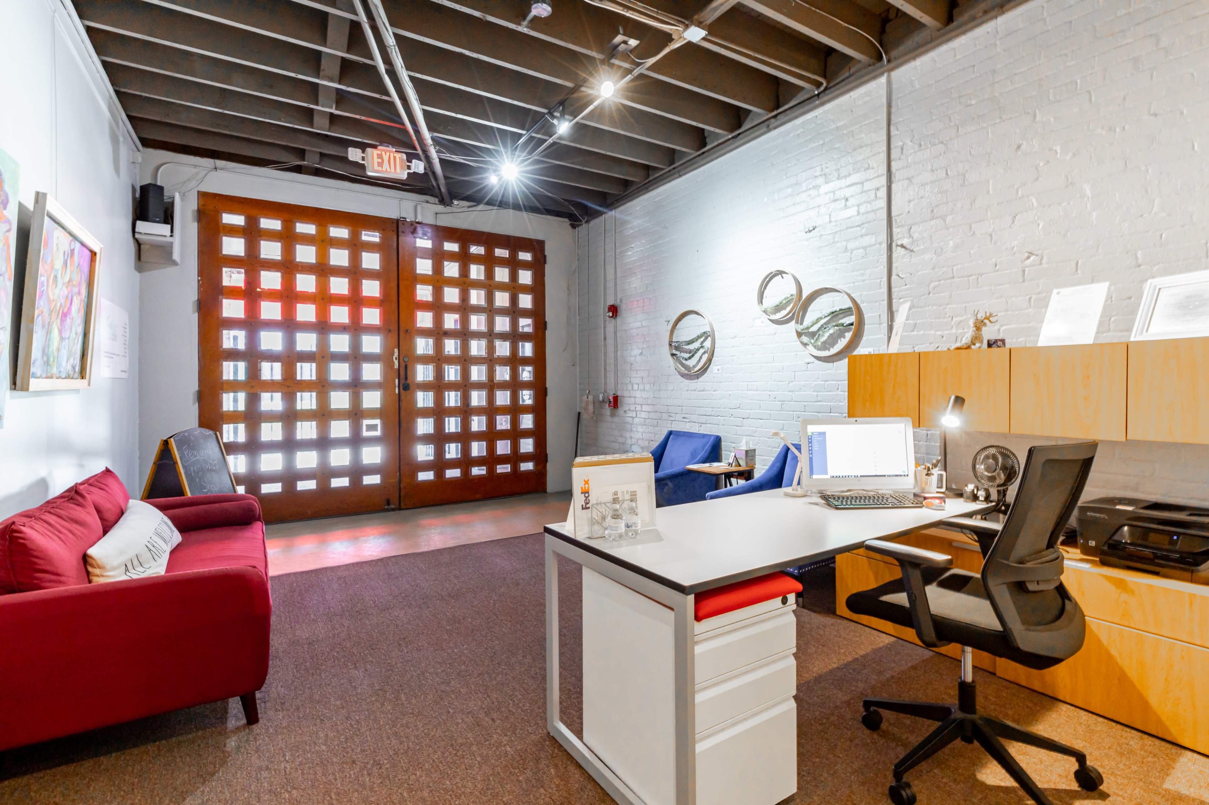 A modern office space featuring a red couch, a wooden desk with a computer, and blue seating, illuminated by overhead lights.