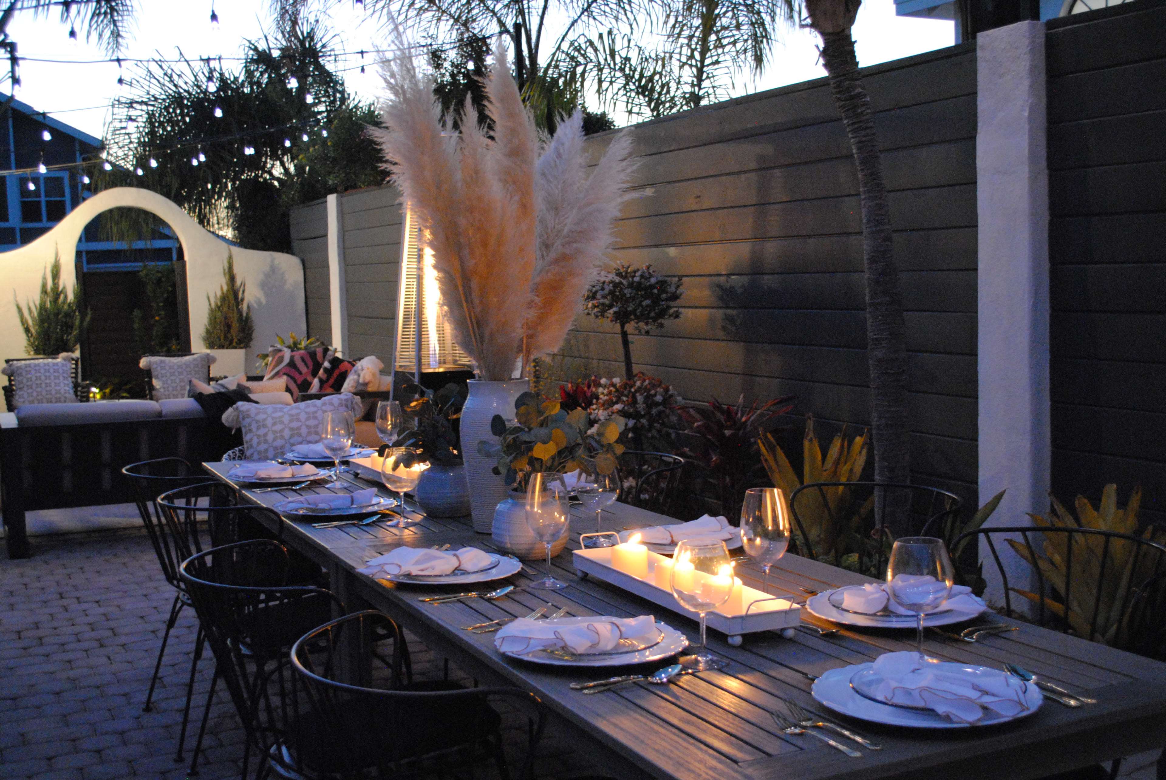 A long outdoor dining table is set with white plates and glasses, surrounded by plants and illuminated by candles at dusk.