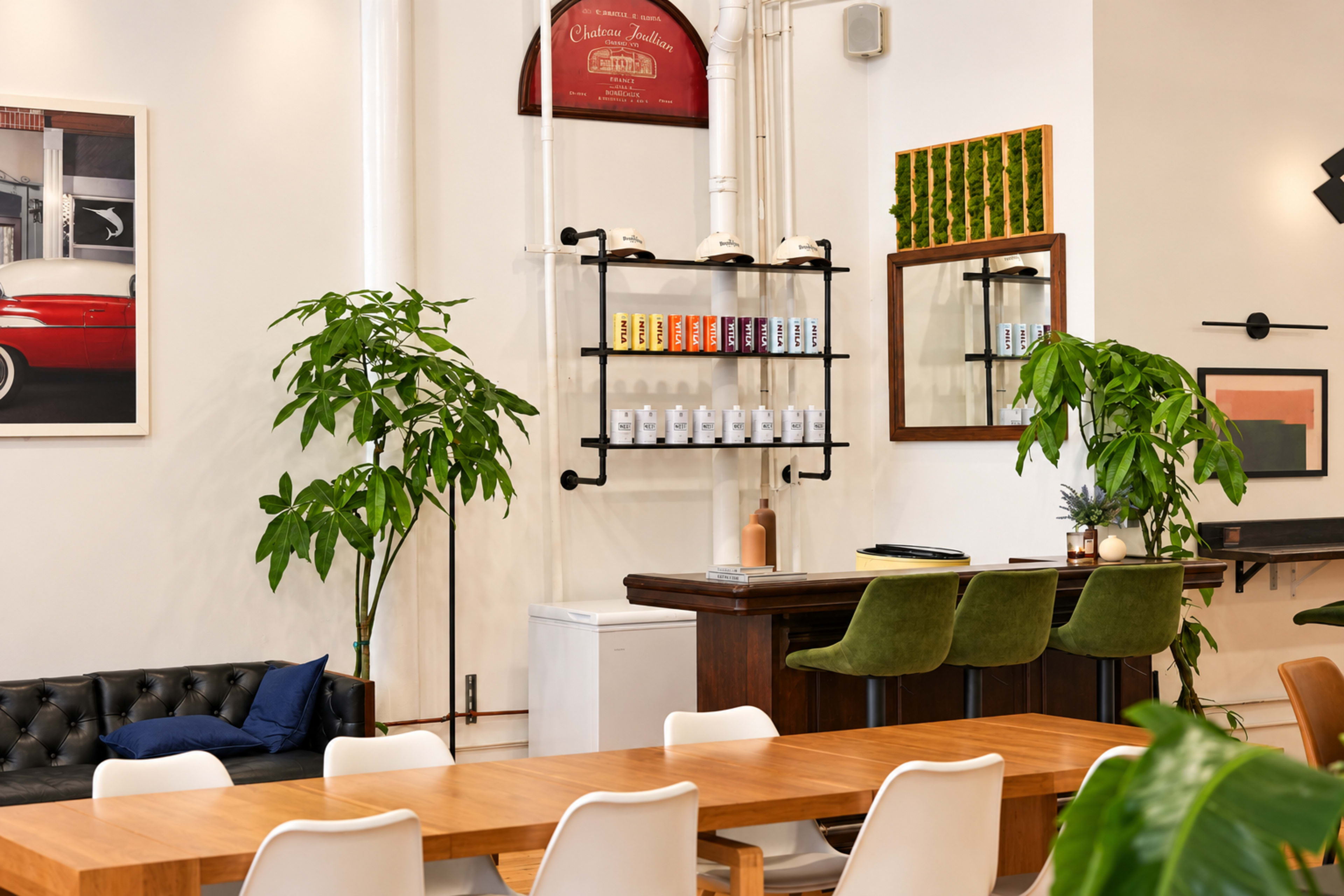 The image shows a modern indoor space featuring a wooden dining table, green upholstered bar stools, a black leather couch, and decorative plants arranged around a bar area with colorful cans displayed on the wall.