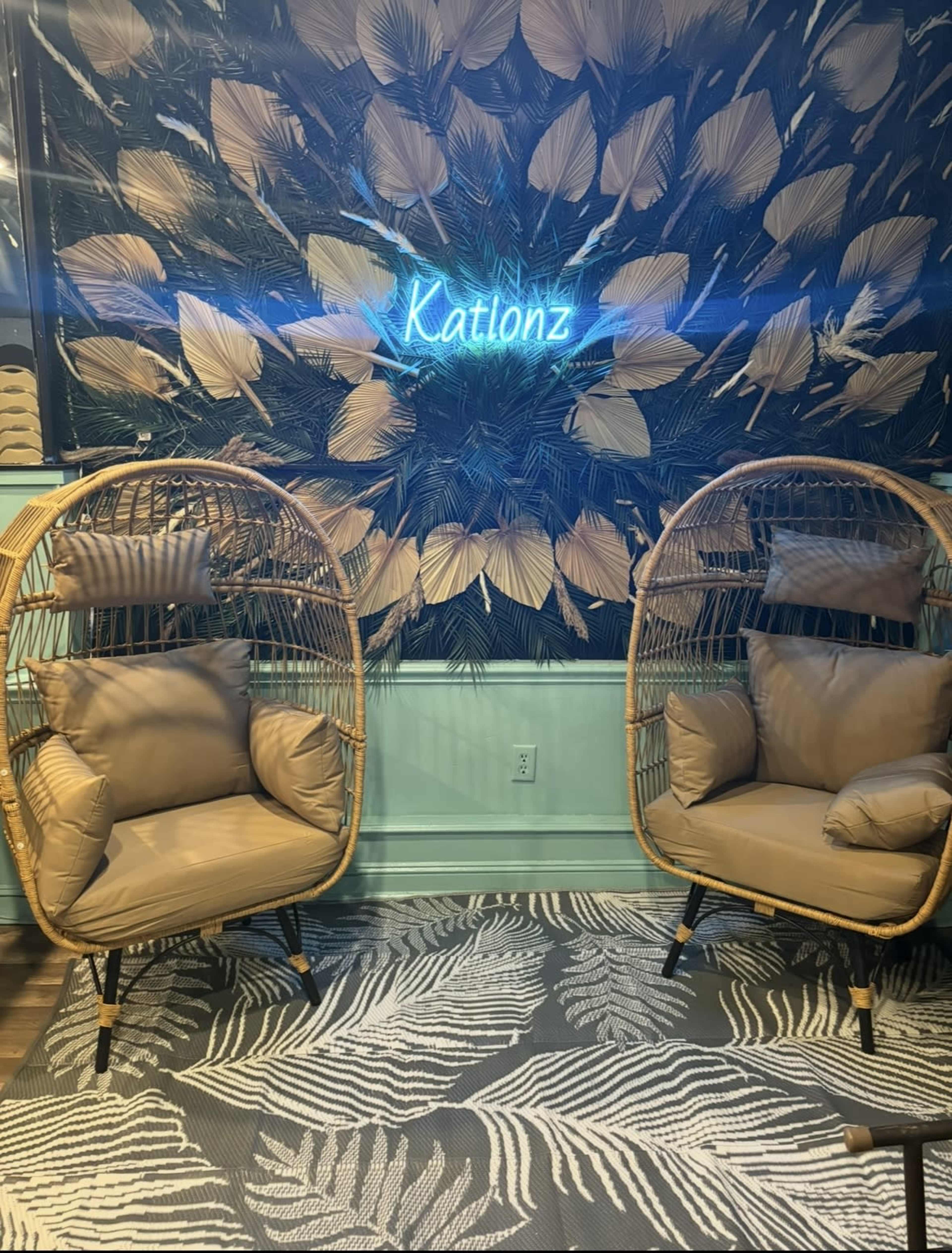 The image features two wicker chairs with cushions positioned in front of a leafy wallpaper and a neon sign that reads "Katlionz."