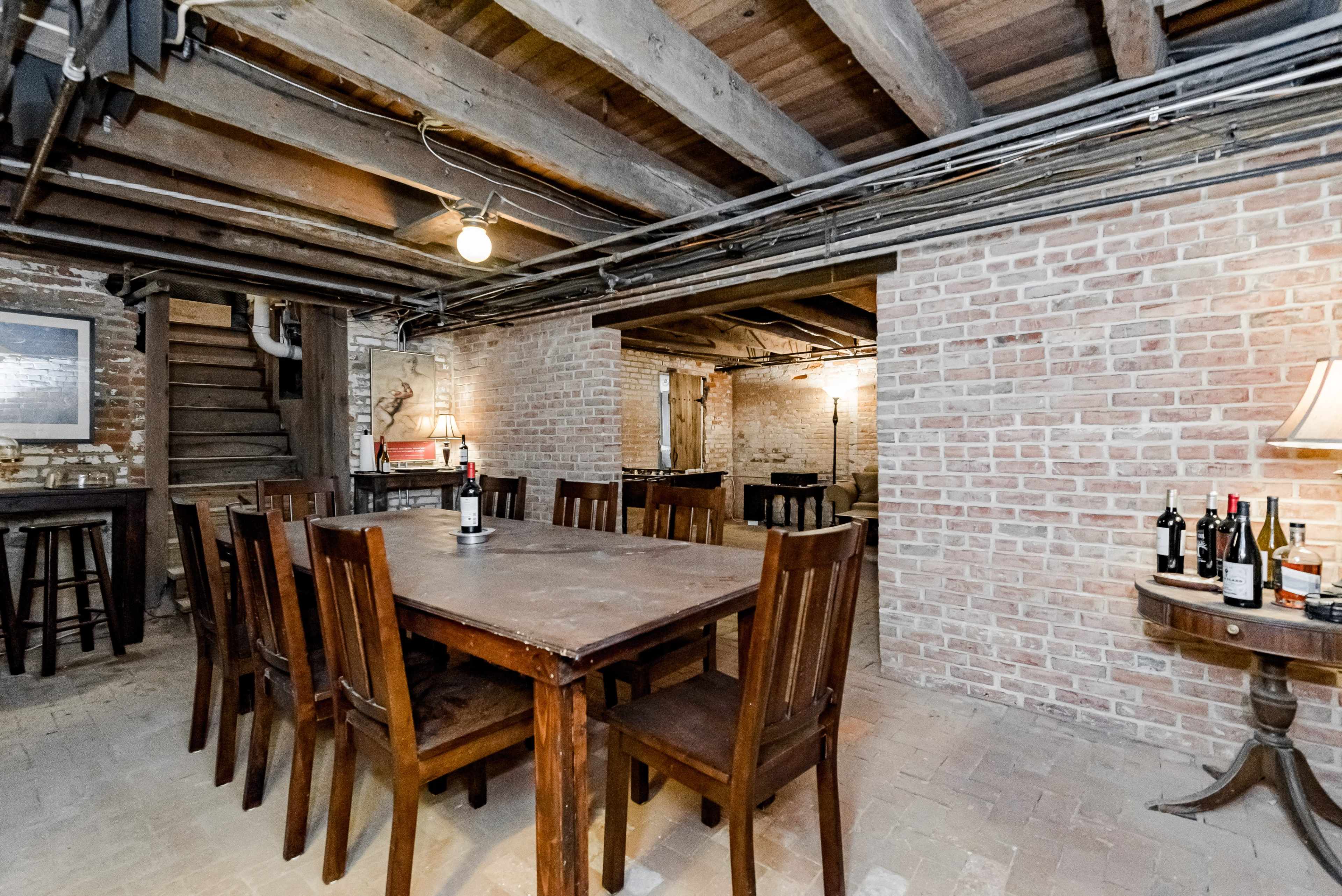 A spacious basement features exposed wooden beams, brick walls, a large wooden dining table surrounded by chairs, and various bottles of wine on a side table.