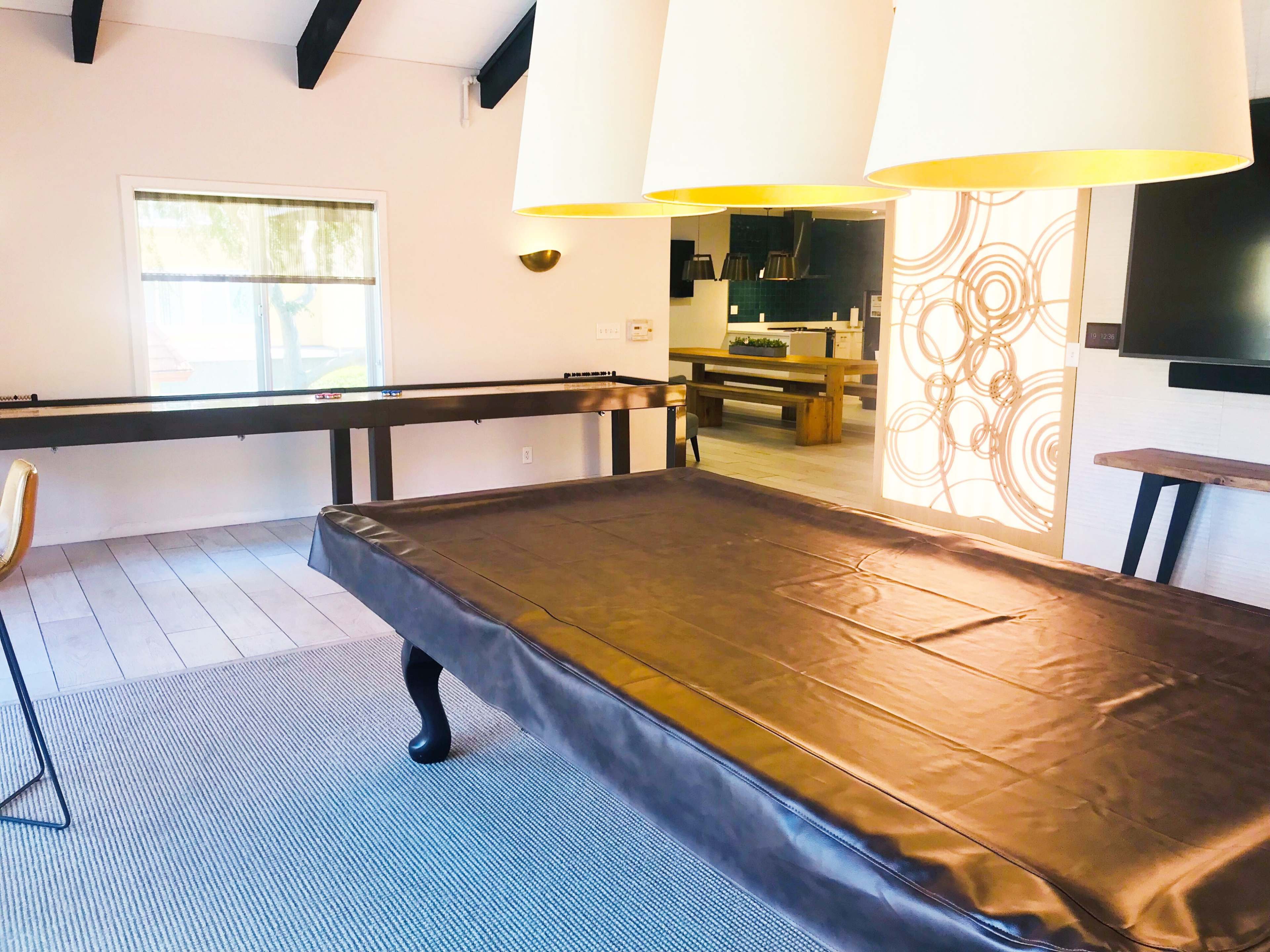 A spacious room with a pool table in the foreground, a dining area in the background, and large pendant lights hanging from the ceiling.
