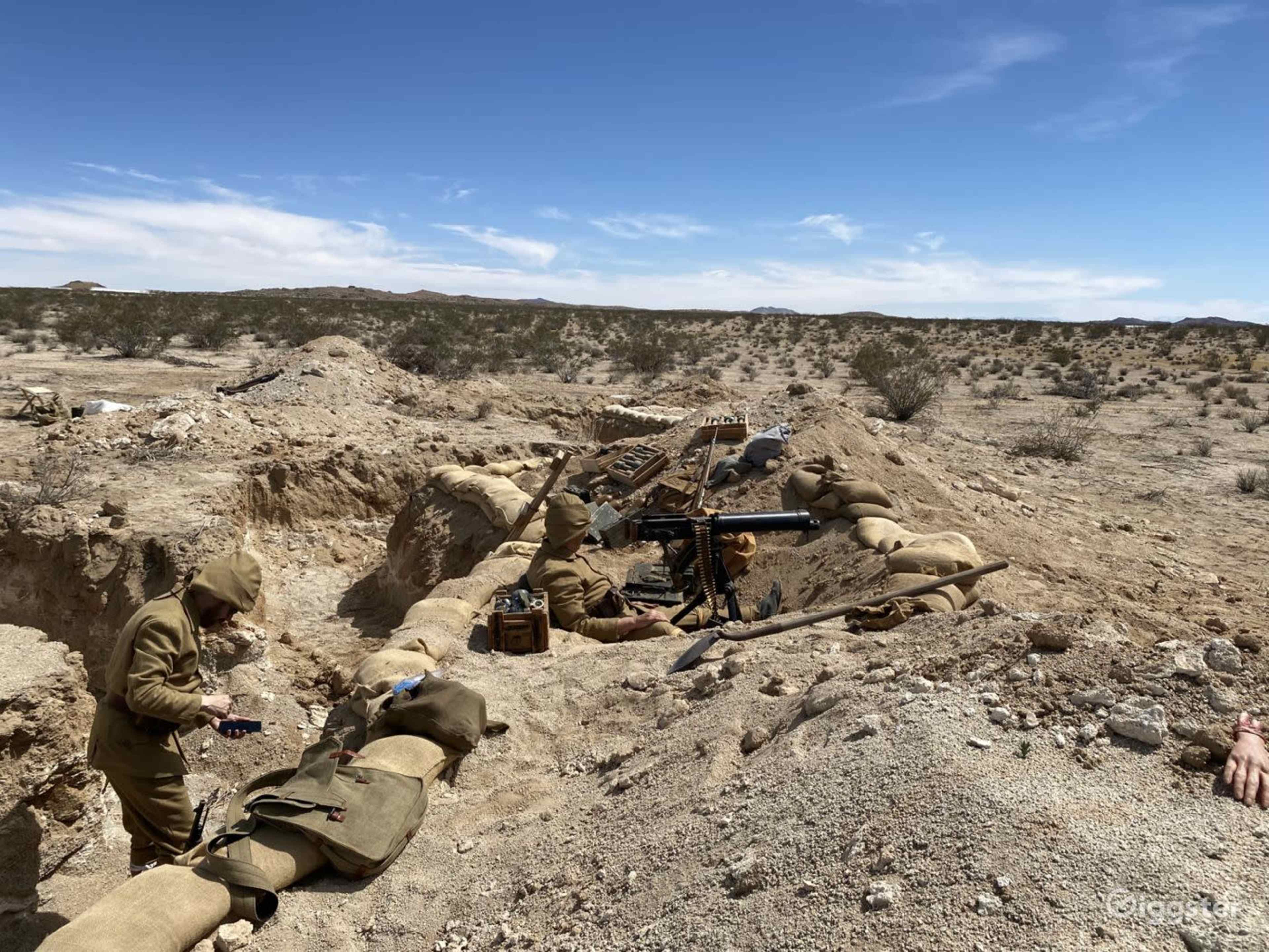 A military trench is set up in a barren landscape, with soldiers in khaki uniforms working near equipment and supplies.