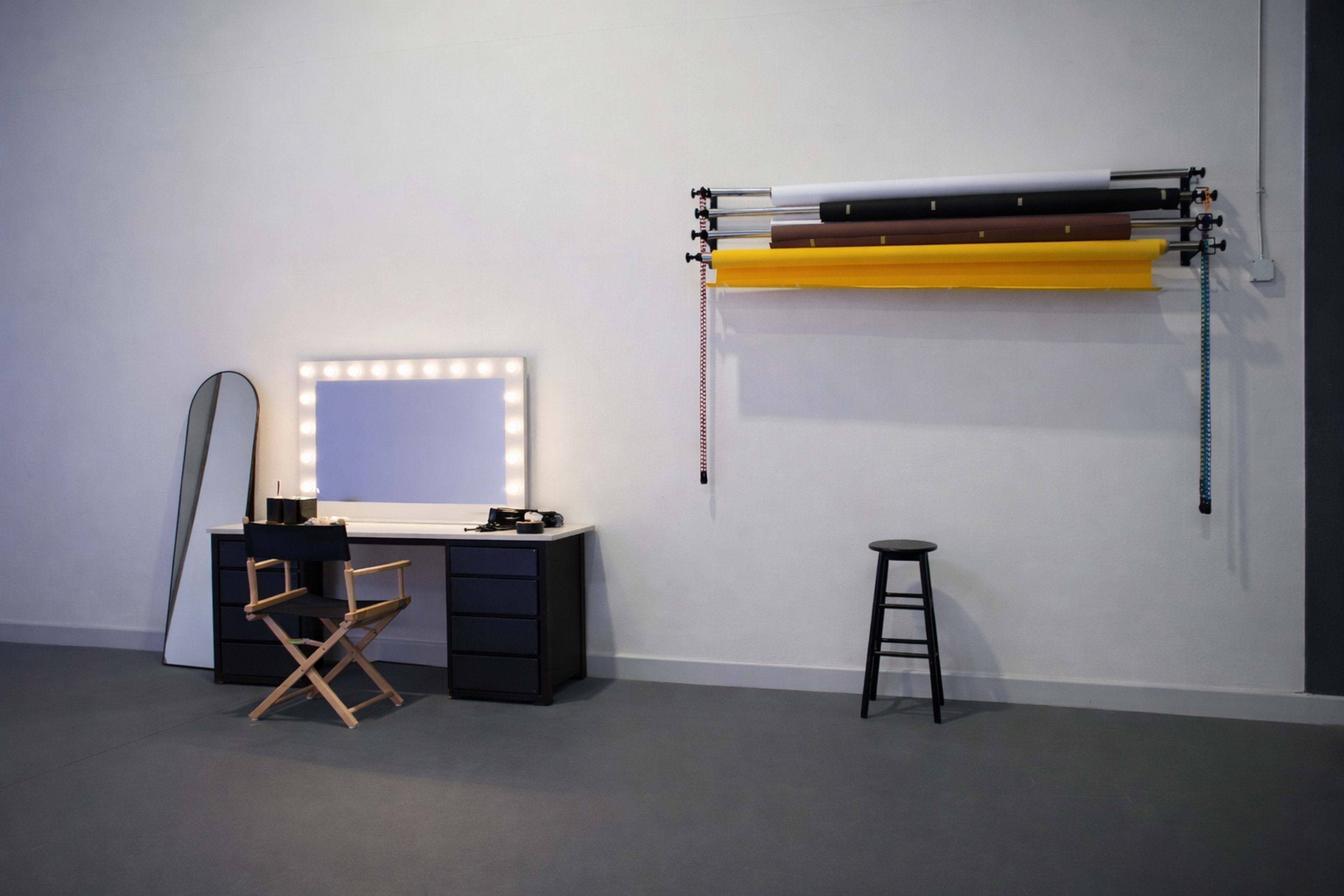 The image shows a minimalistic studio space featuring a dressing table with lights, a director's chair, a stool, and rolls of colored backdrops mounted on the wall.