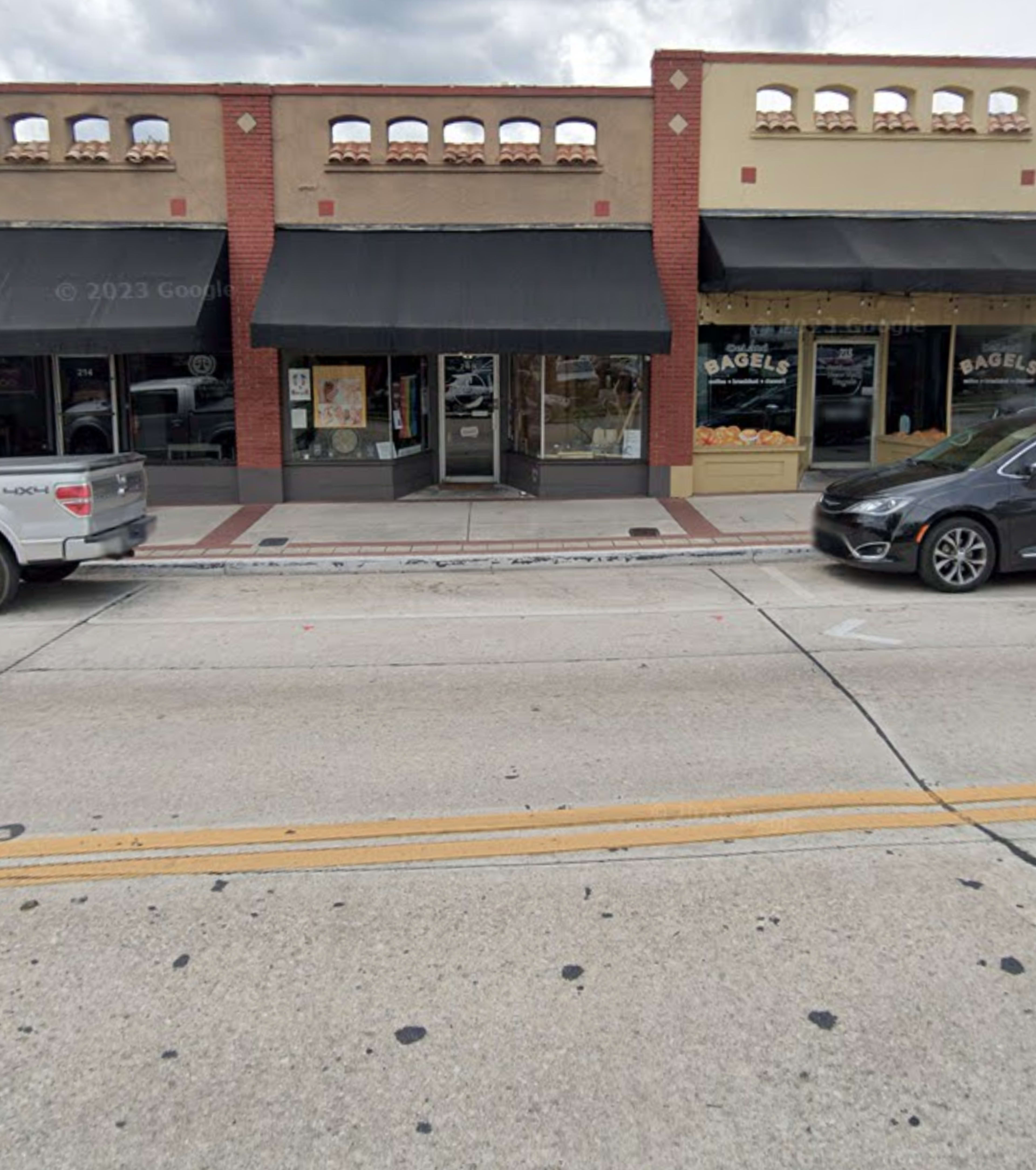 A street view of a row of storefronts, including a bagel shop, with parked cars along the roadway.