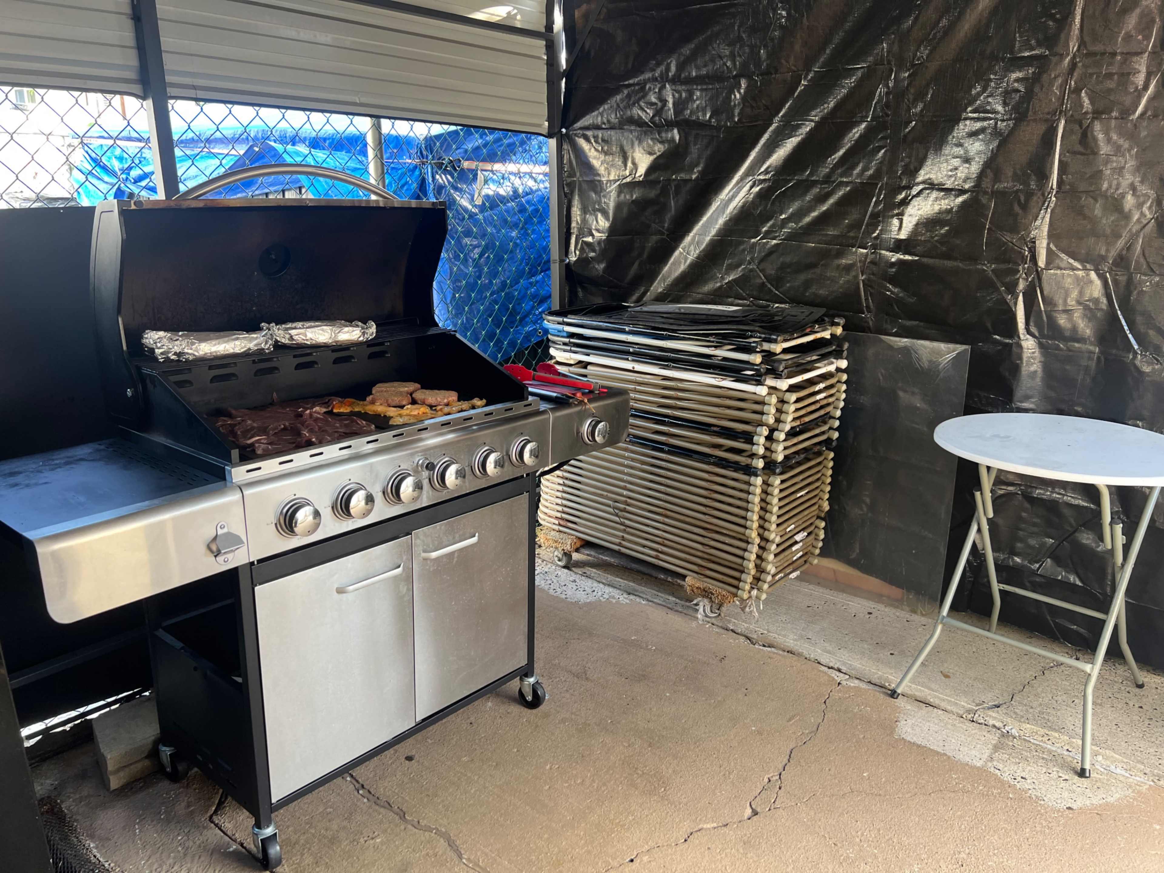 A grill with various meats and vegetables is positioned next to a stack of folding tables in a covered outdoor area.